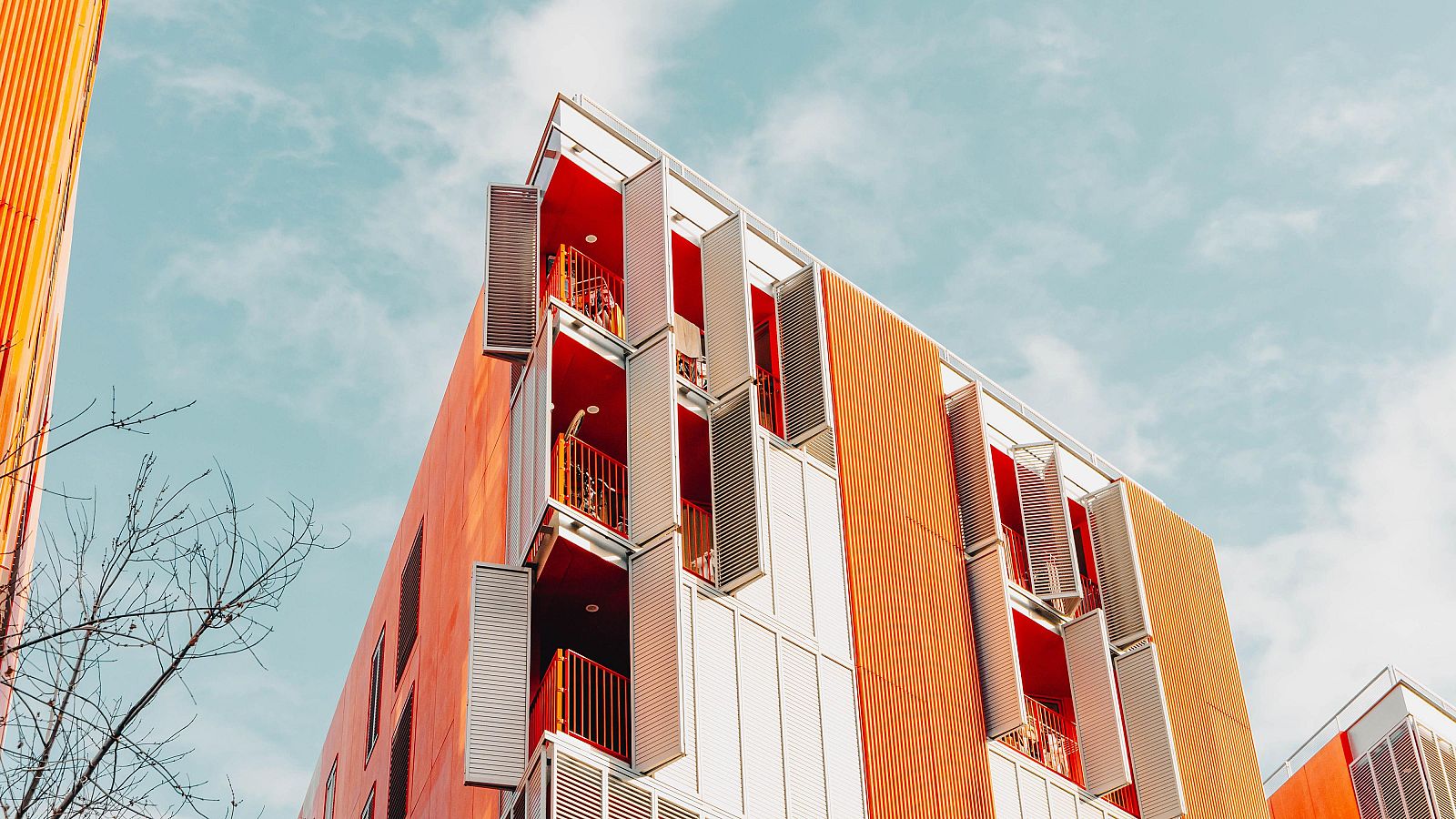 Edificio de viviendas moderno, con fachada naranja y blanca, balcones con persianas metálicas y detalles naranjas. Perspectiva desde abajo, cielo azul de fondo.