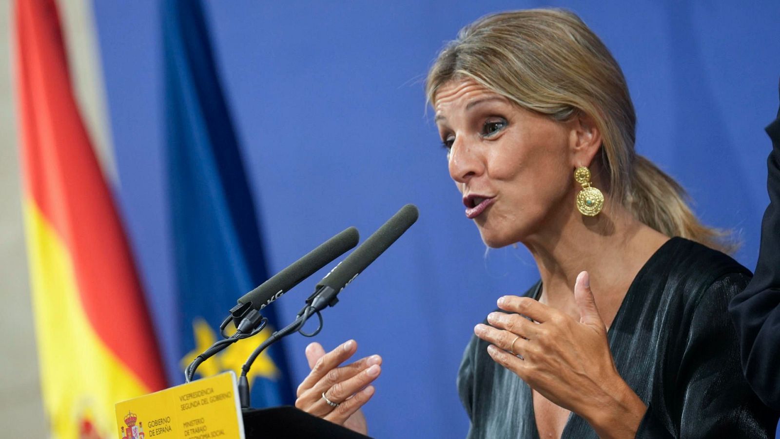 Política hablando en atril con dos micrófonos, blusa negra y pendientes dorados.  Banderas de España y la UE al fondo. Gestos enérgicos.
