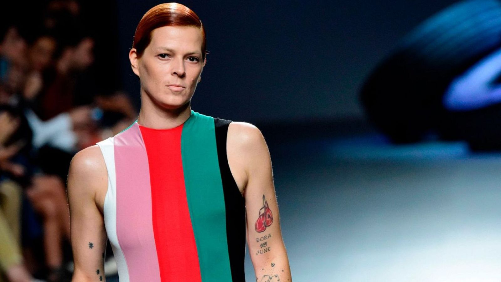 Bimba Bosé, con vestido multicolor y tatuajes visibles, posa con expresión seria.  Pelo corto rojizo hacia atrás.  Fondo desenfocado sugiere un evento.