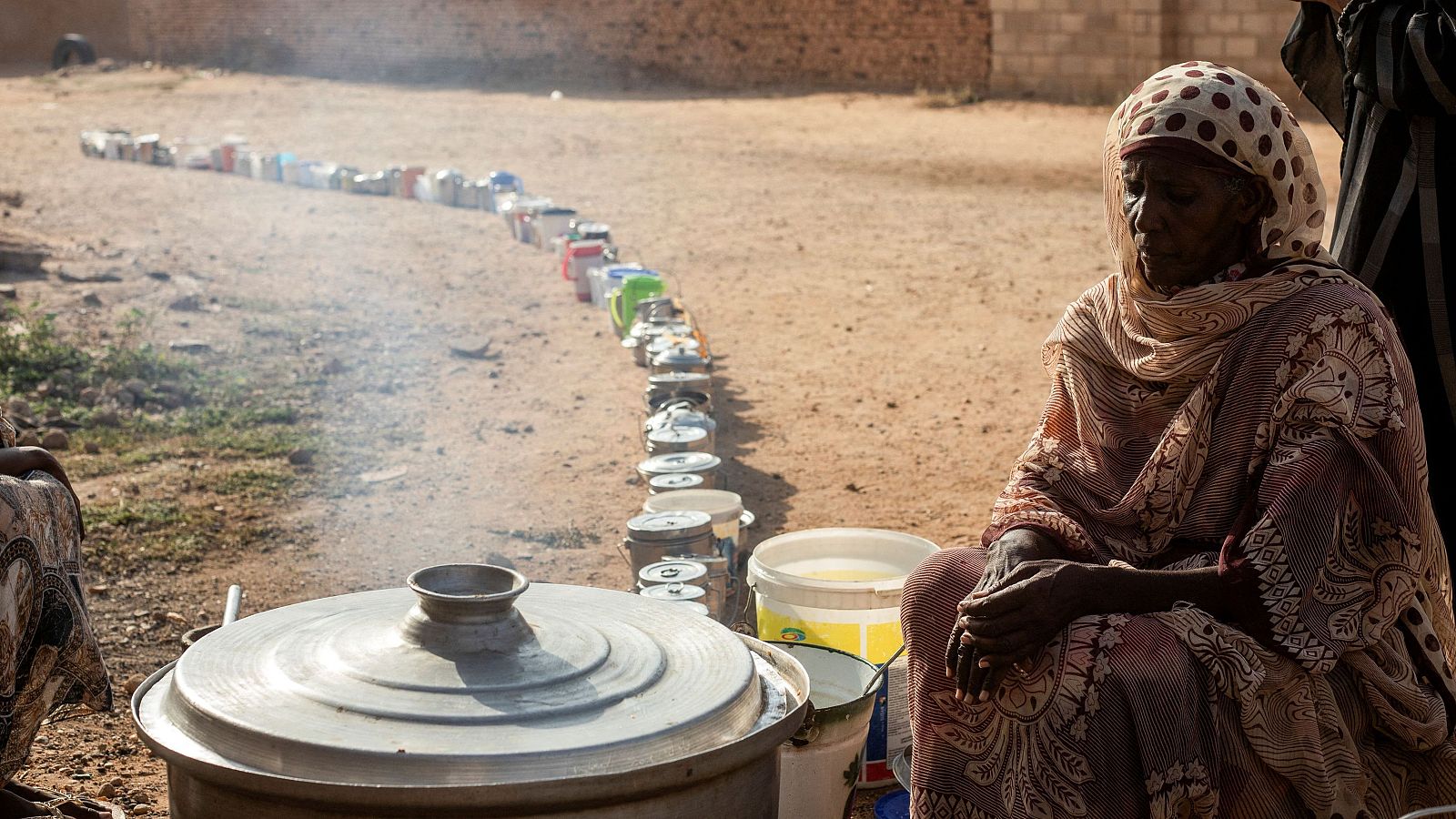 Mujer cocinando en una olla grande; vapor se eleva;  fila de recipientes esperan ser llenados.