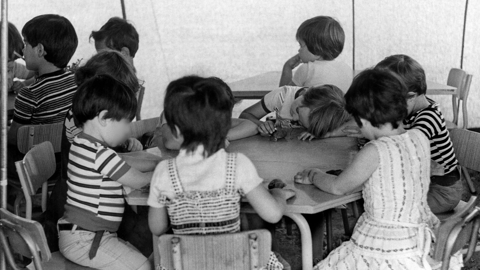 Niños de entre 4 y 7 años sentados en mesas de madera dentro de una tienda, algunos con camisetas de rayas.  Ambiente infantil, posiblemente educativo o lúdico.