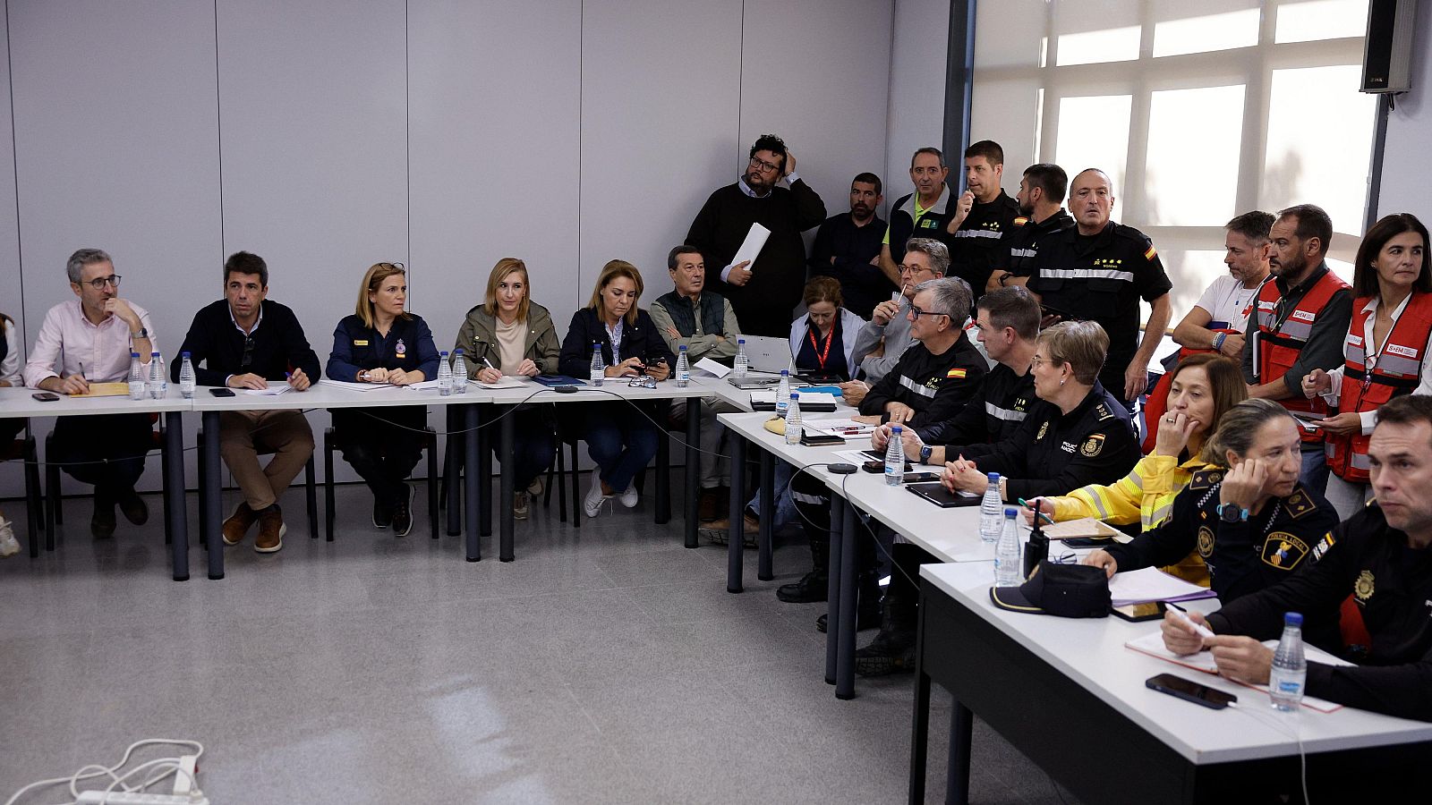 Coordinación de emergencias en una sala de reuniones.  Participantes con uniformes y ropa civil usan ordenadores y dispositivos de comunicación.
