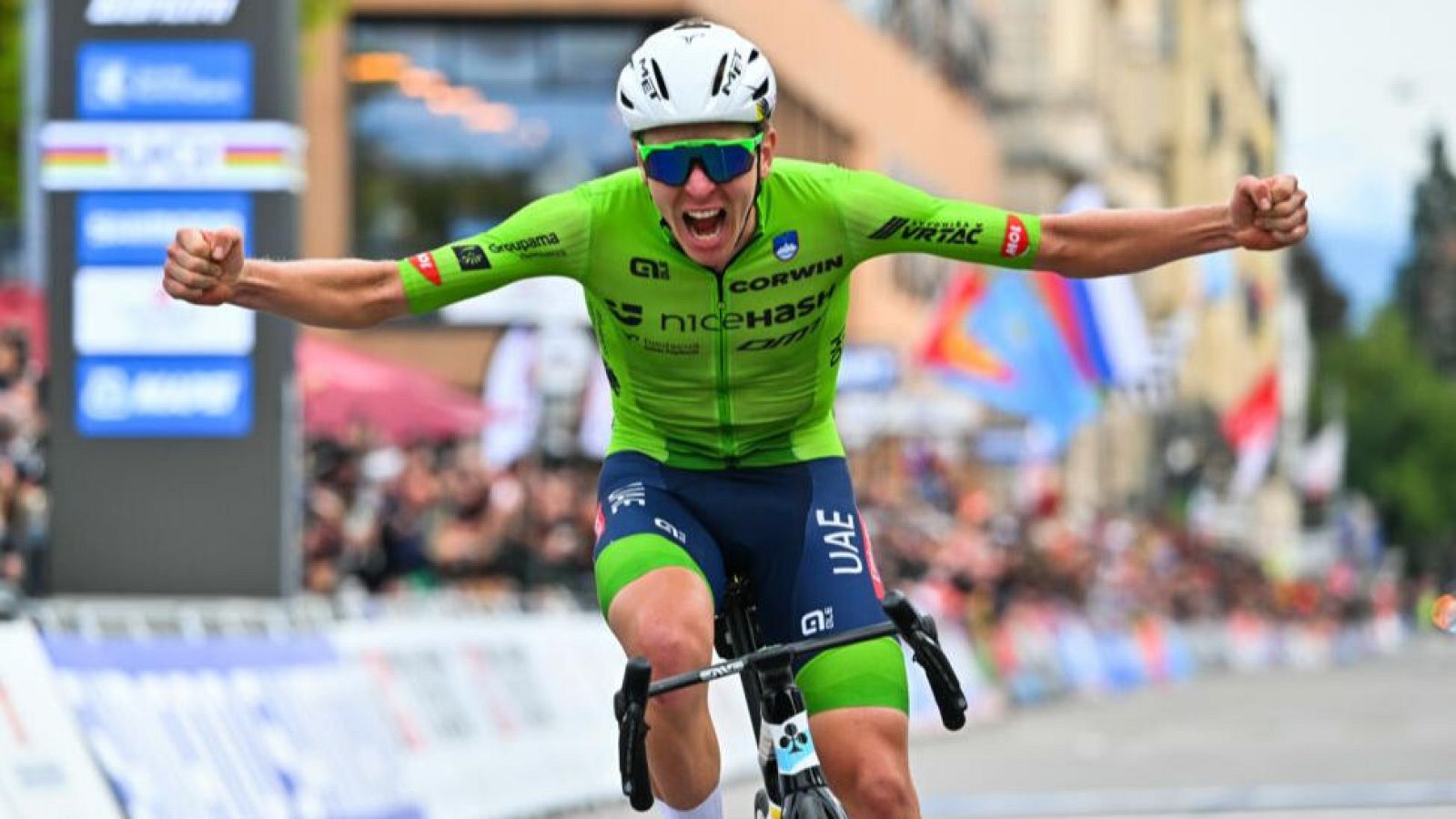 Ciclista en maillot verde con logos, celebra victoria alzando los brazos.  Gesto de euforia, fondo con público y banderas desenfocadas.