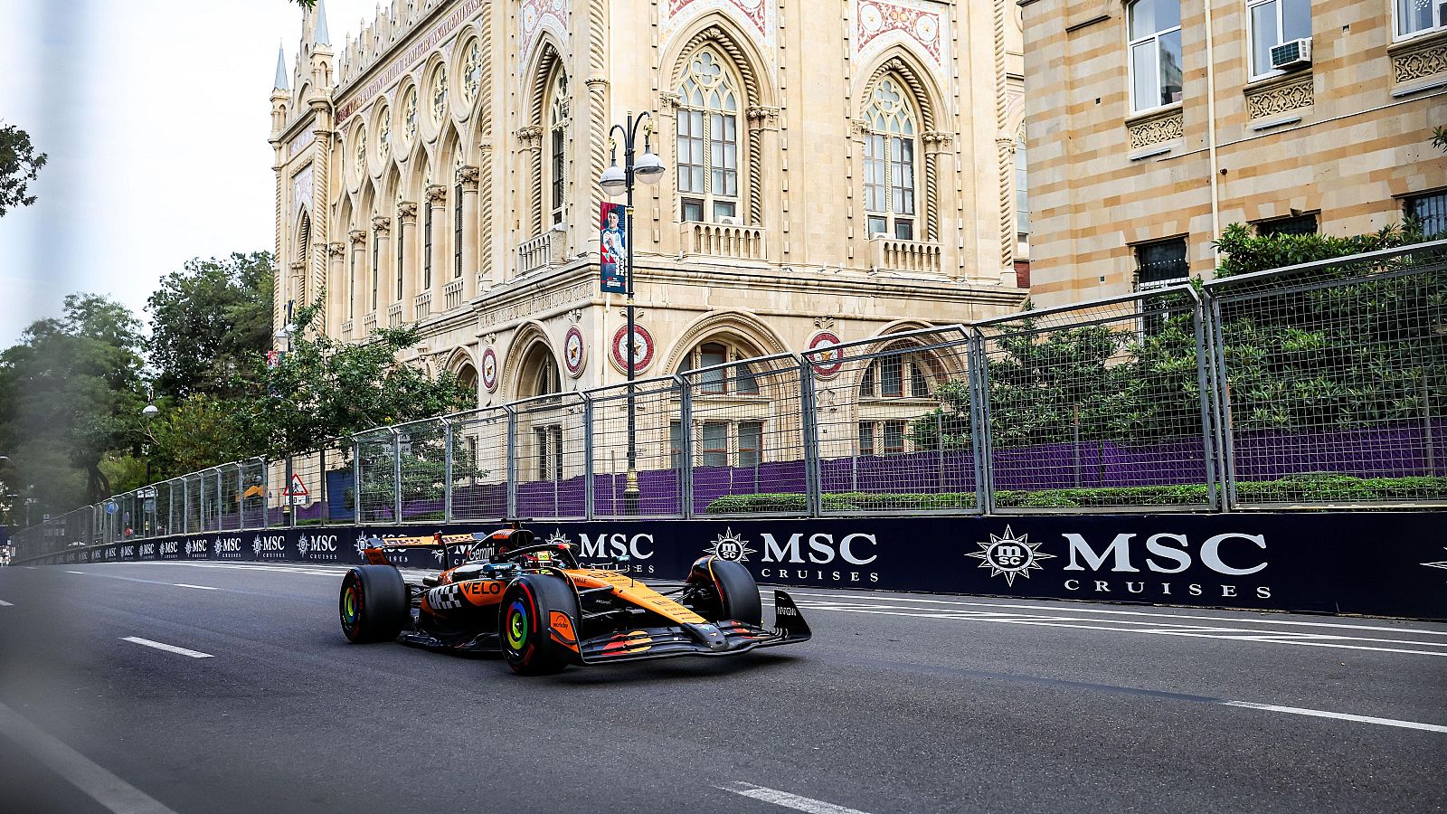 Monoplaza McLaren en acción durante entrenamientos libres en circuito urbano.  El coche, de colores naranja y azul, se desplaza junto a un edificio clásico y vallas con publicidad.