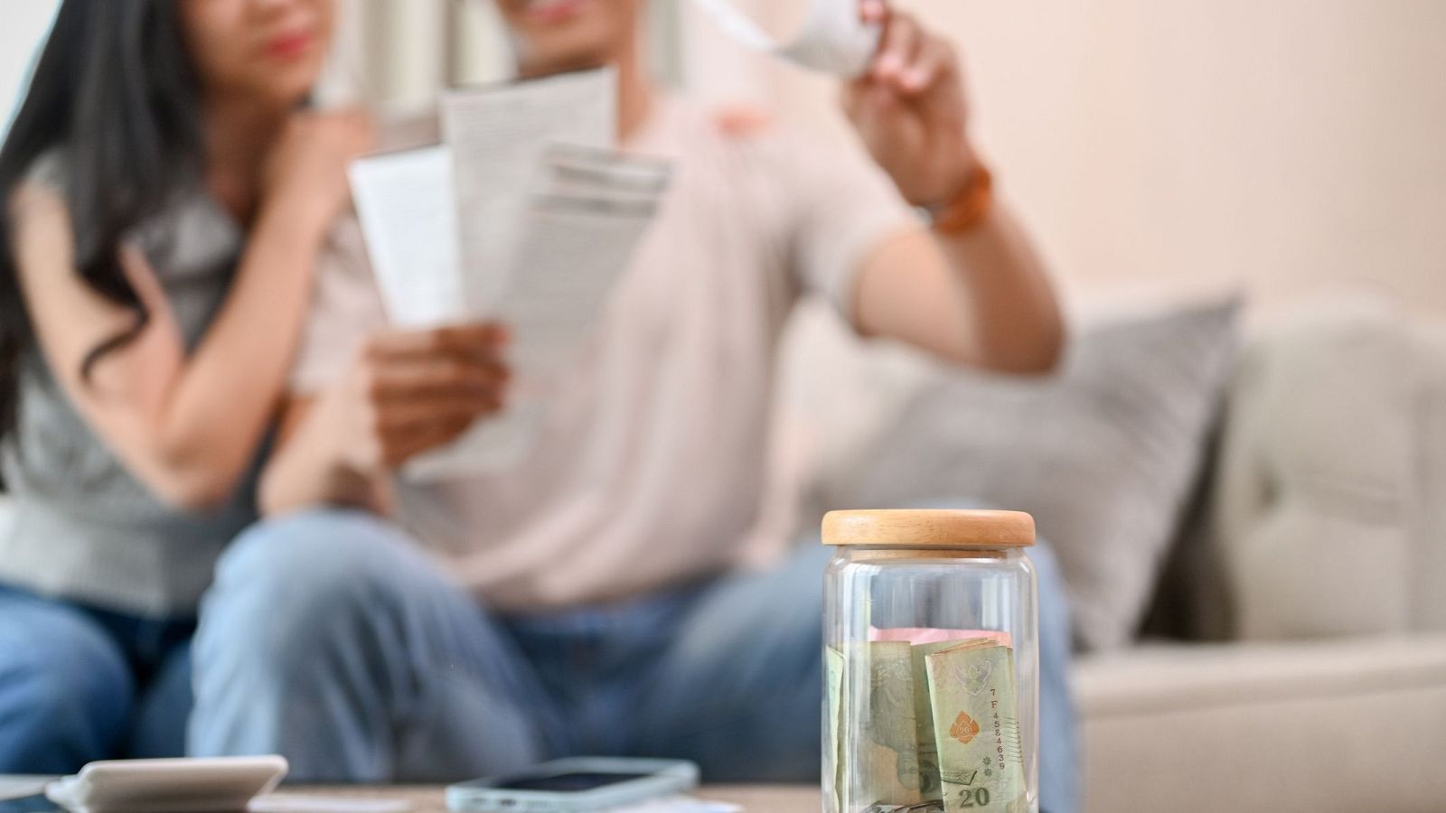 Frasco de vidrio con ahorros en efectivo, junto a una pareja que parece estresada revisando papeles.  Imagen que refleja preocupaciones económicas.
