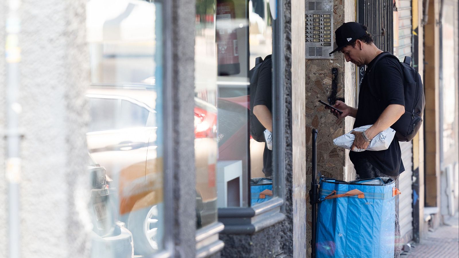 Repartidor con paquete y móvil en mano, junto a una bolsa de reparto.  Su vestimenta es oscura y lleva gorra.
