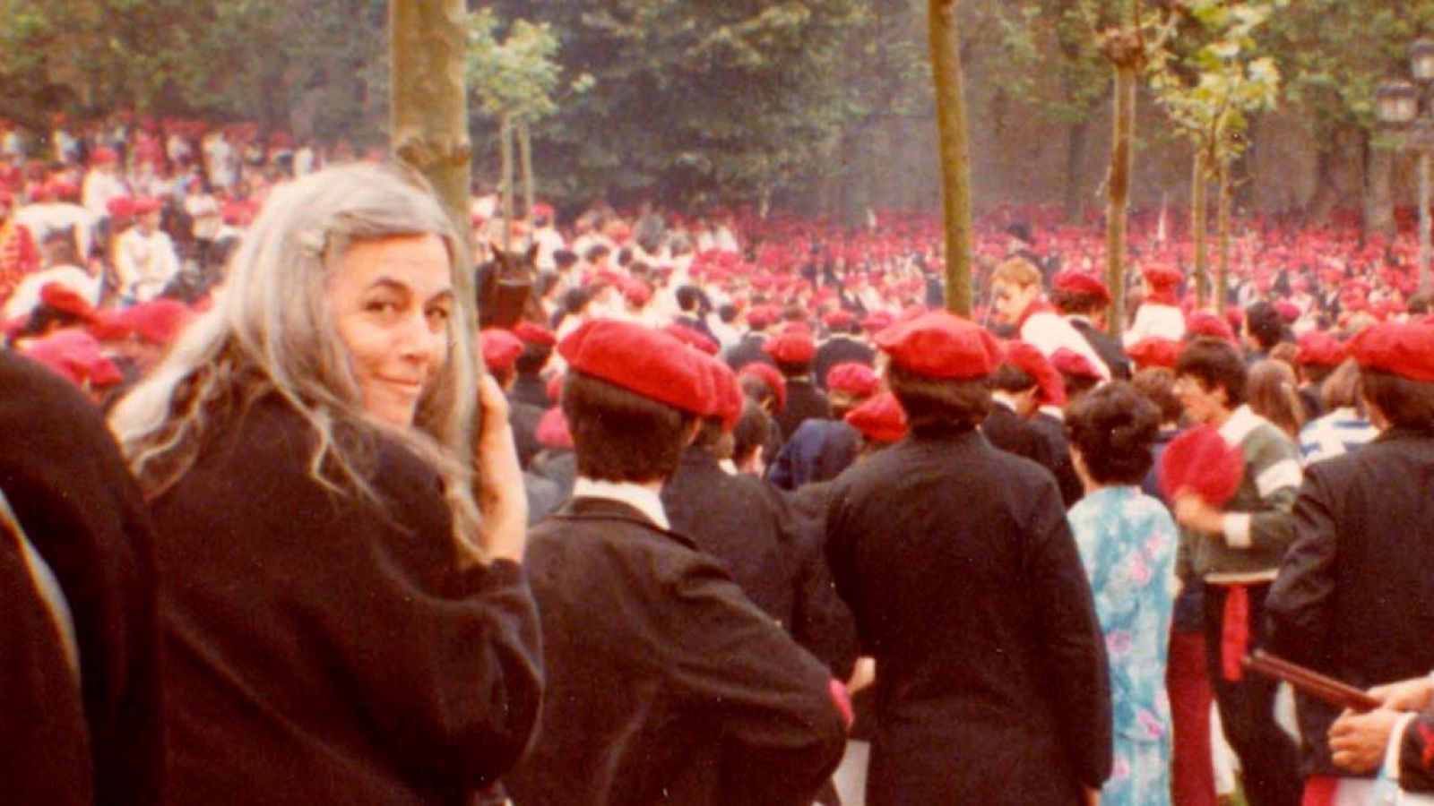 Primer plano de Carmen Martín Gaite sonriendo a cámara; viste ropa oscura.  Fondo con multitud de personas con boinas rojas en exterior.