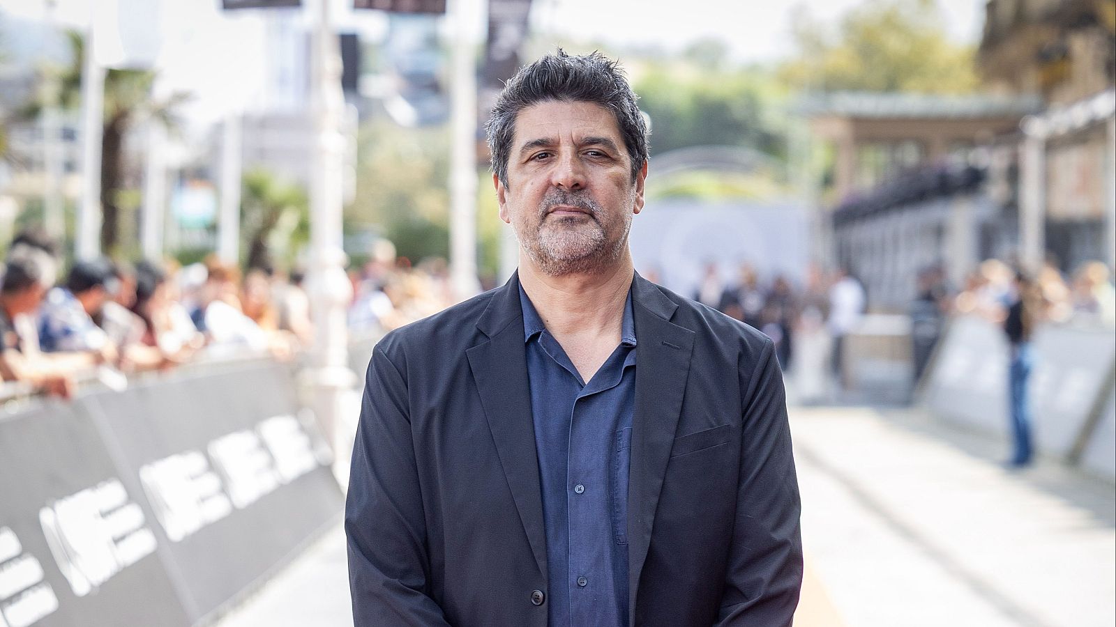 Primer plano de Cesc Gay, director de cine, mirando a cámara. Lleva chaqueta oscura y camisa azul. Fondo con multitud y alfombra roja.