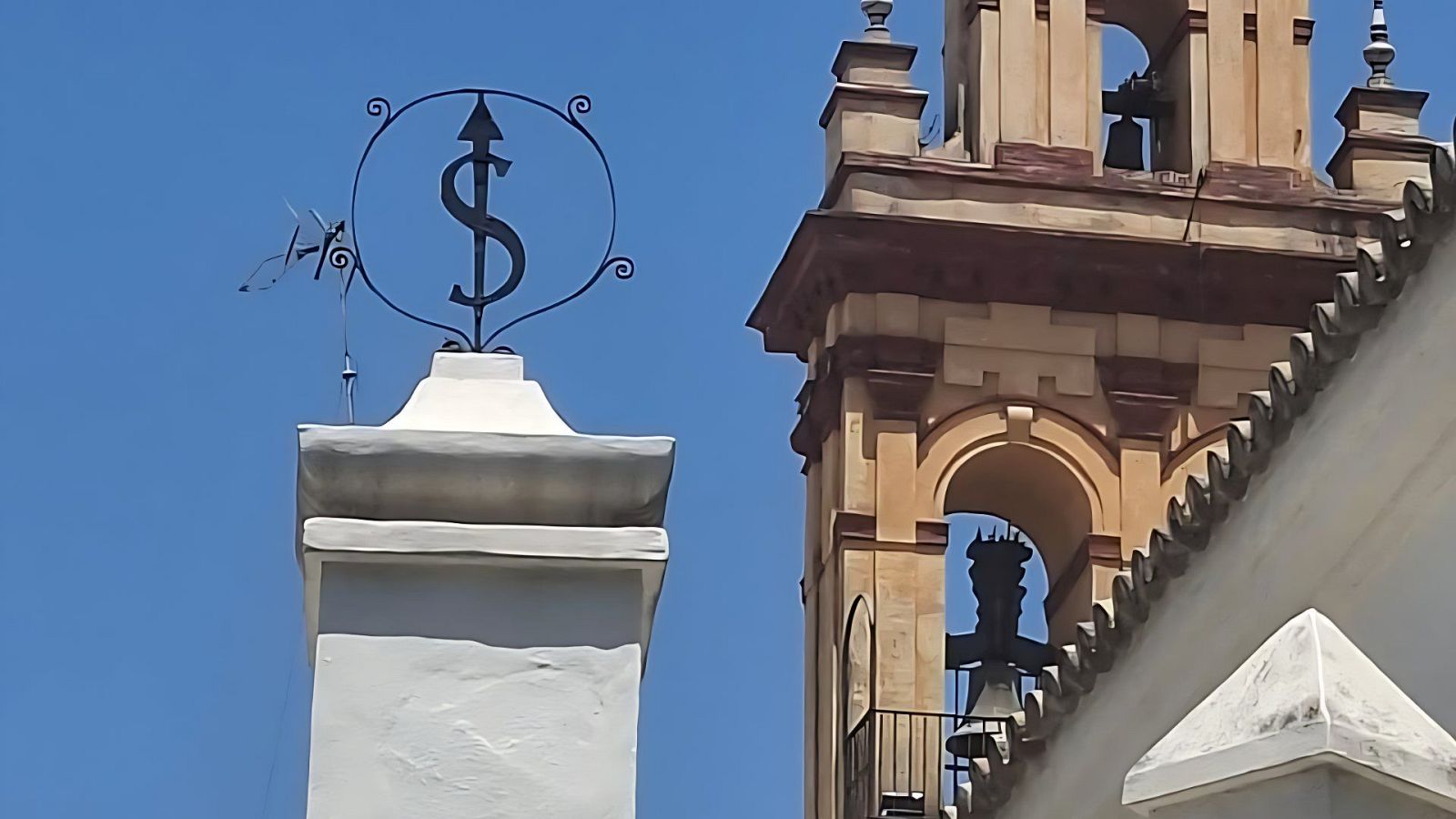 Símbolo de hierro forjado en forma de 'S' con flecha, dentro de un círculo, sobre columna blanca.  Parte de campanario beige visible.  Cielo azul claro.