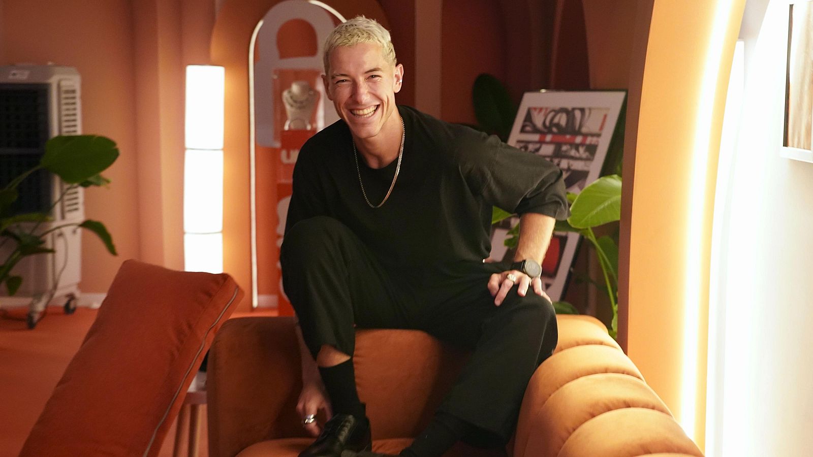Hombre con ropa negra, sentado en un sofá anaranjado, sonríe a cámara. Lleva anillos, reloj y cadena. Fondo con planta y aparato de aire acondicionado.