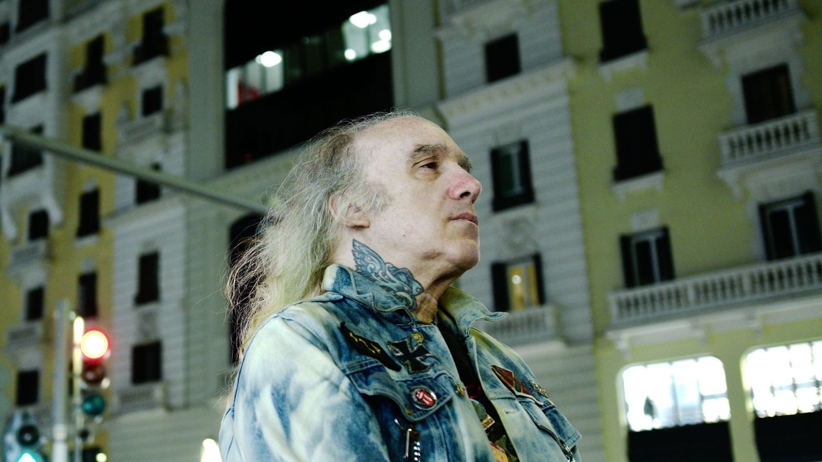 Un hombre con cabello largo y canoso, con una chaqueta vaquera y un tatuaje en el cuello, se encuentra de pie en la calle, con un edificio alto y un semáforo como telón de fondo.