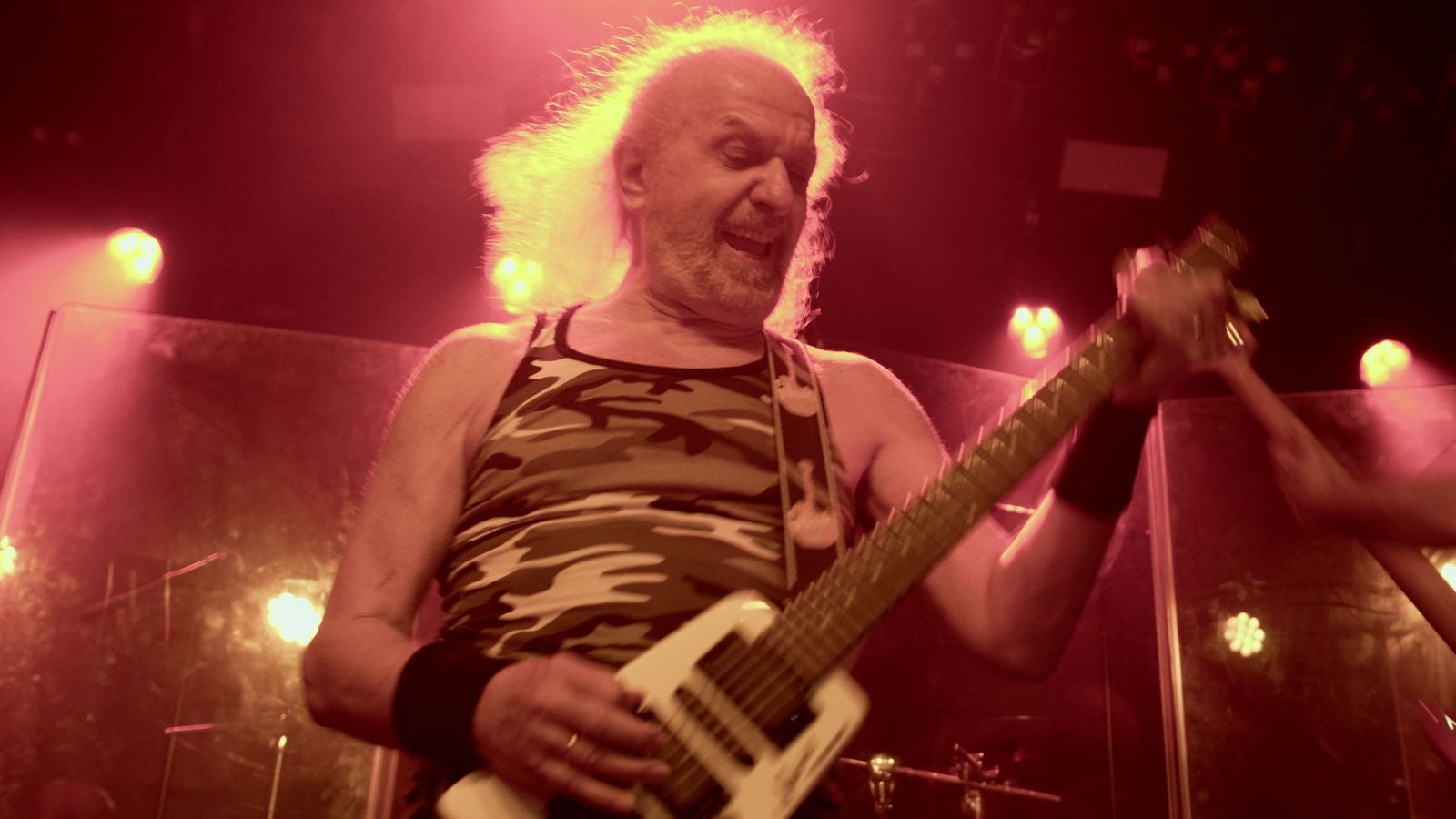 Un guitarrista con camiseta de camuflaje y muñequeras negras toca una guitarra eléctrica en un escenario iluminado con luces rojas, posiblemente durante una actuación musical.