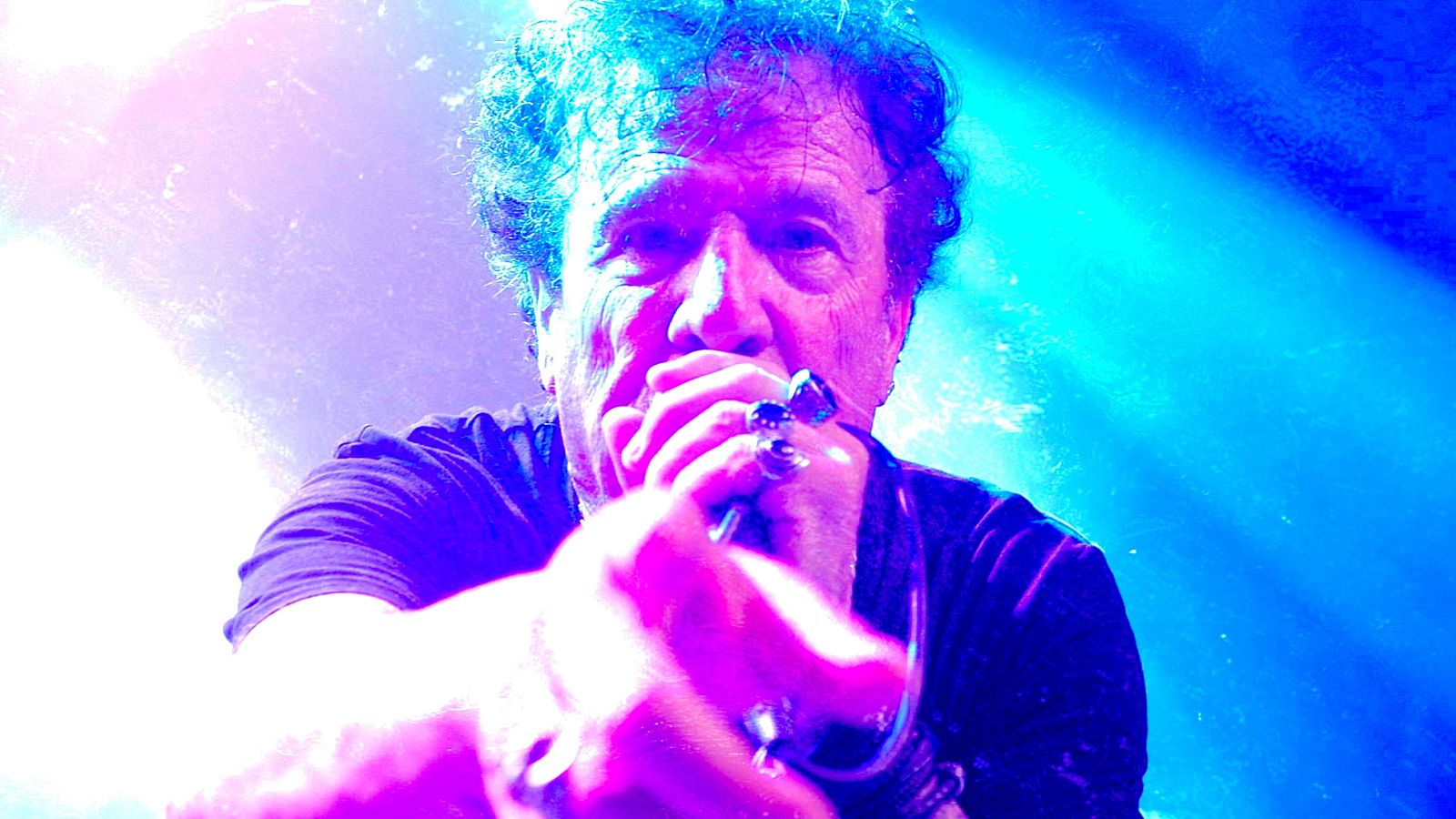 Un hombre con cabello rizado y oscuro, posiblemente un músico, canta o grita con intensidad a un micrófono, con una camiseta negra y anillos, en un ambiente con iluminación azul y morada.
