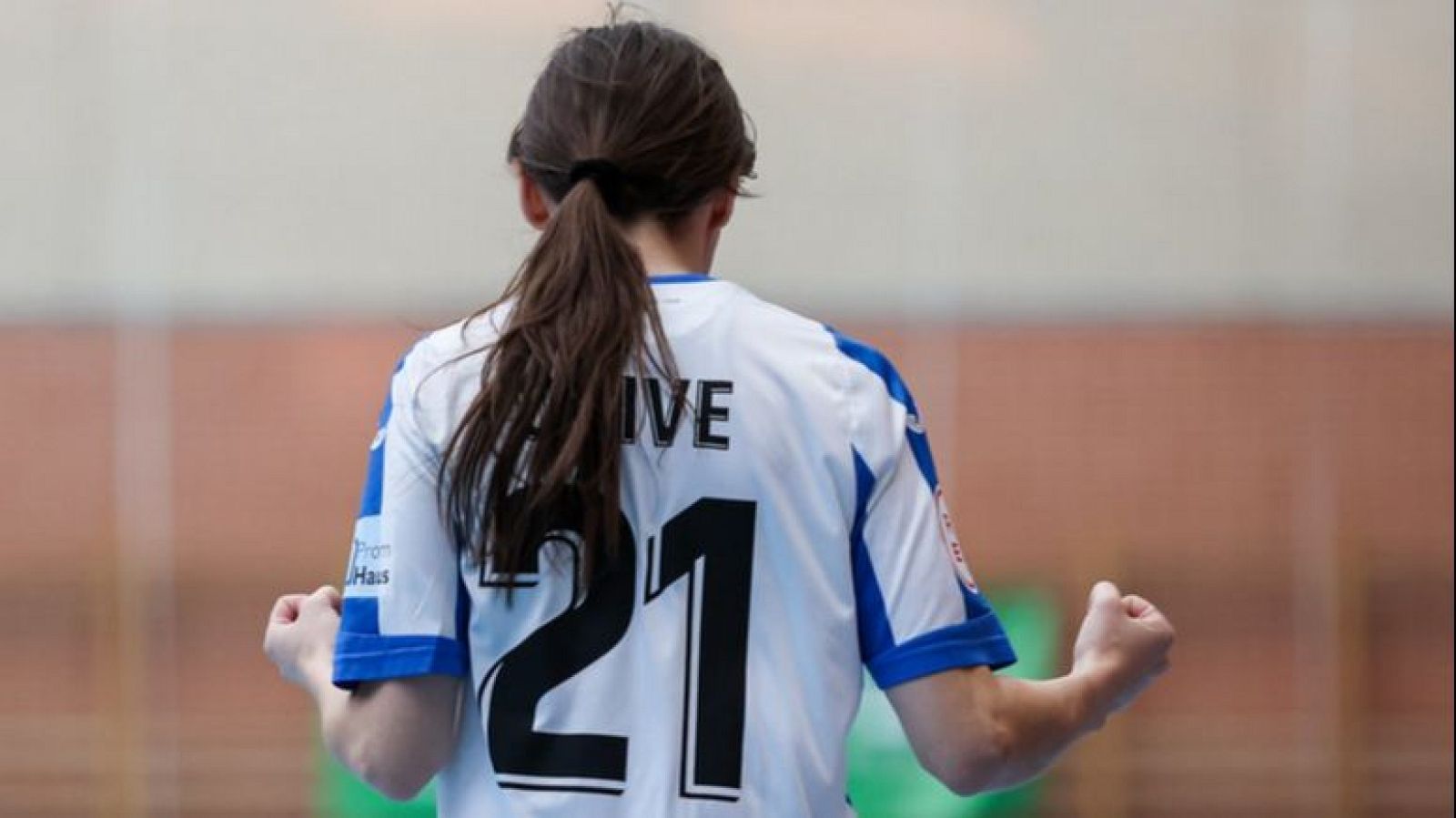 Una jugadora de fútbol sala, con el pelo recogido en una coleta, celebra un gol con los puños cerrados. Lleva una camiseta deportiva blanca con detalles azules y el nombre y número del dorsal visibles en la espalda.