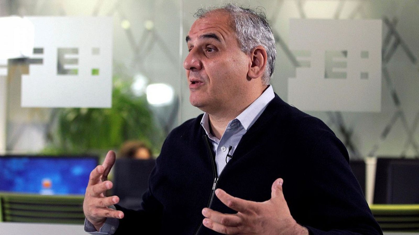 Empresario de mediana edad con camisa blanca y chaqueta oscura gesticulando en una oficina moderna con logo "FE" visible.