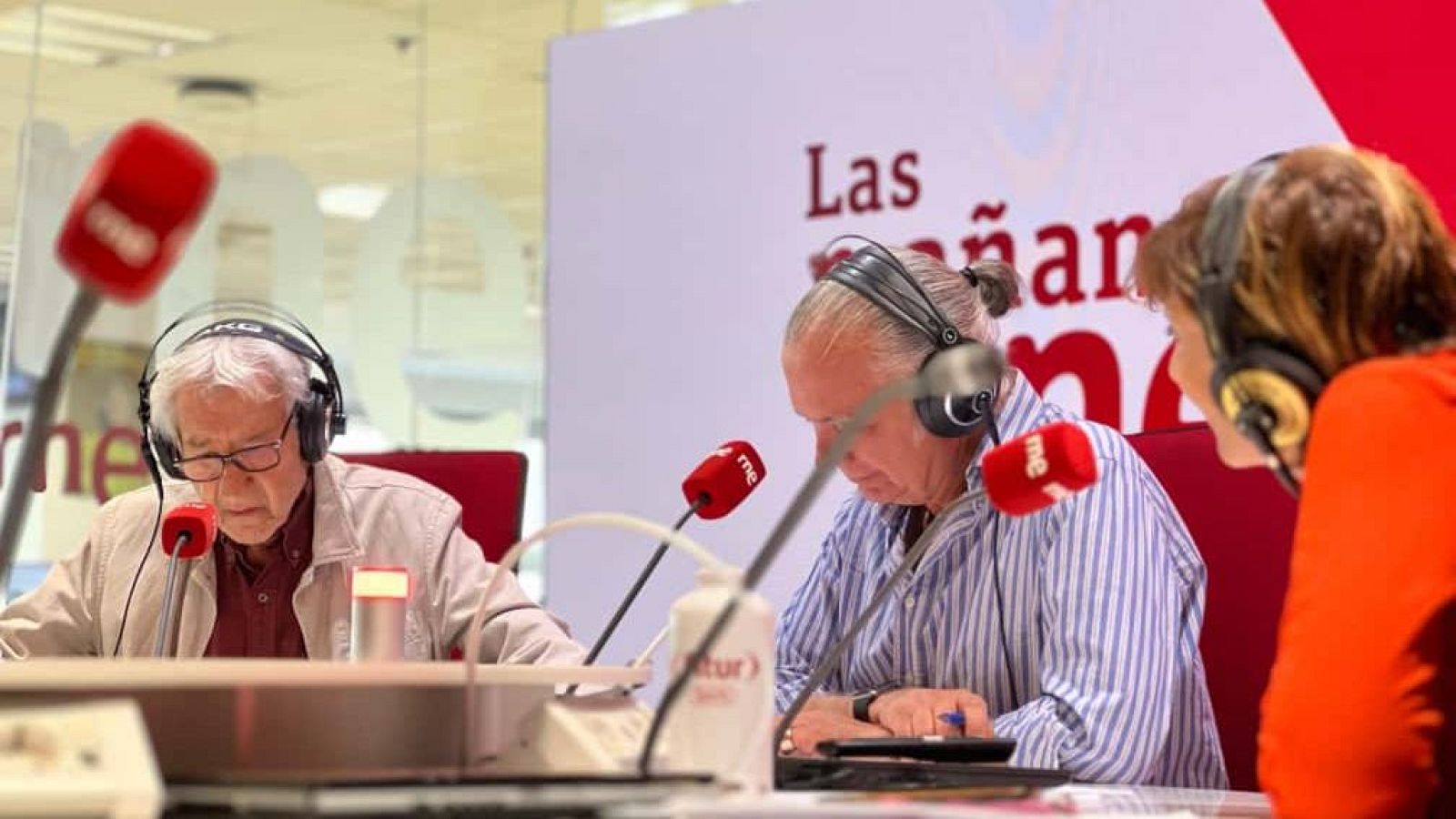 En un estudio de radio, tres personas con auriculares están sentadas frente a micrófonos. Se distingue a un hombre mayor con gafas y chaqueta clara, otro con coleta y camisa de rayas, y una persona con camisa naranja.