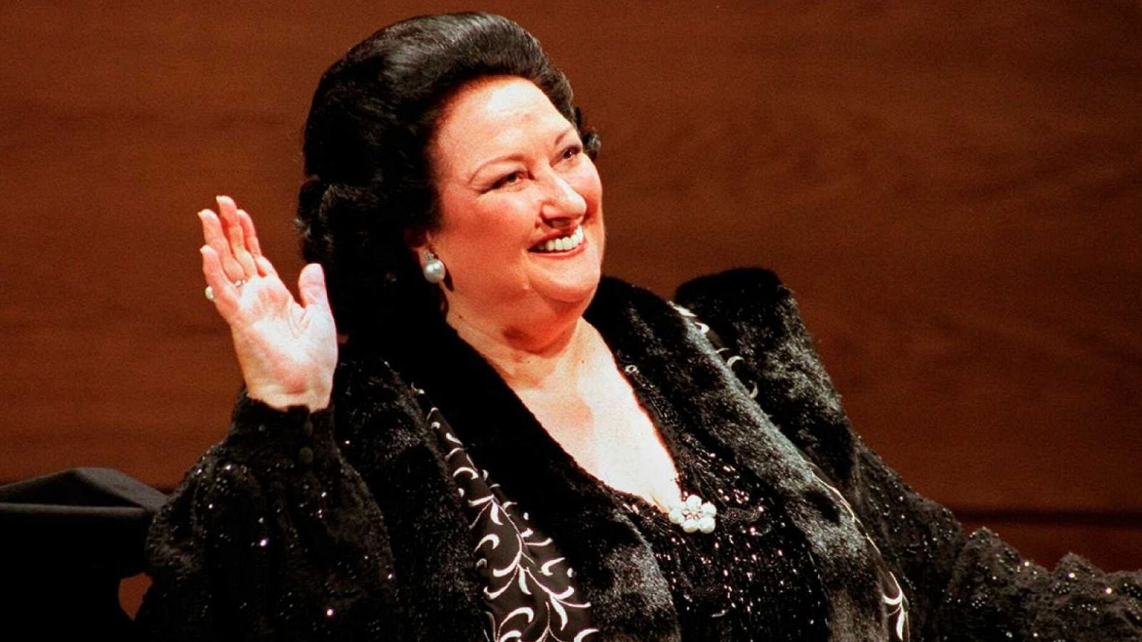 Montserrat Caballé, con el cabello peinado hacia atrás y pendientes de perlas, sonríe y saluda. Viste un abrigo de piel negro con detalles brillantes y un broche floral, sobre una blusa estampada en blanco y negro.