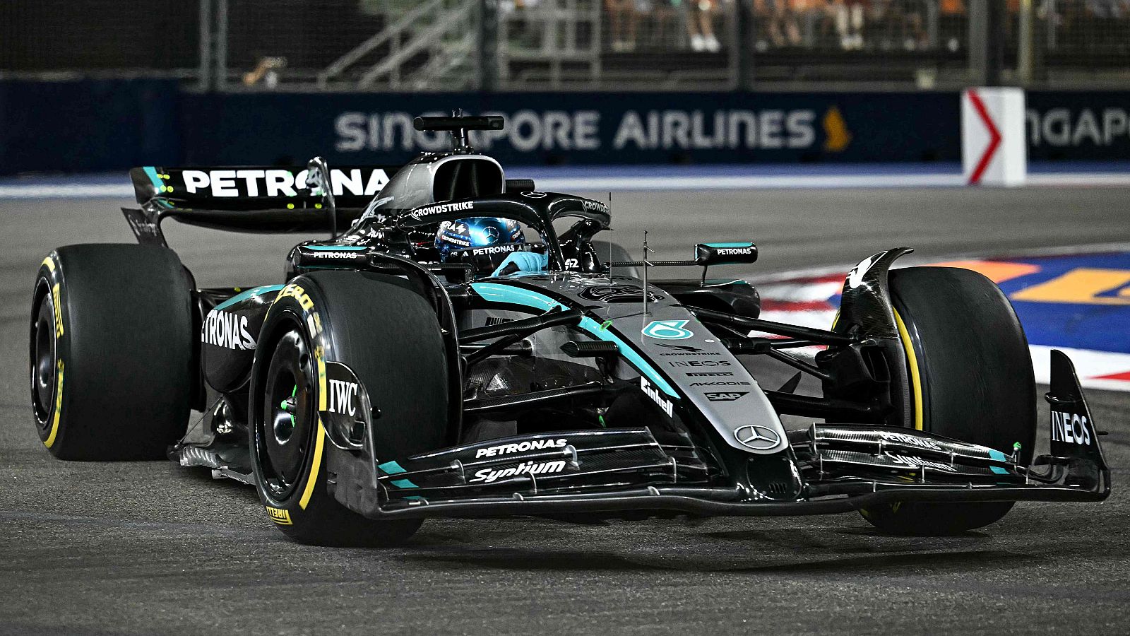 Un monoplaza de Fórmula 1 Mercedes, con detalles en turquesa, compite en una carrera nocturna. Se aprecian logotipos de patrocinadores y publicidad en el fondo, con una pista bien iluminada.