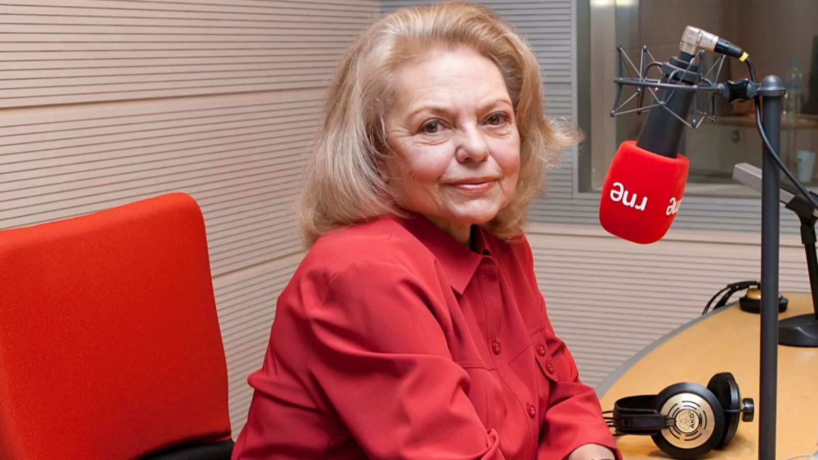 En un estudio de radio, una mujer con cabello rubio y camisa roja está sentada frente a un micrófono con el logo de una emisora. Delante de ella, sobre una mesa, hay unos auriculares.