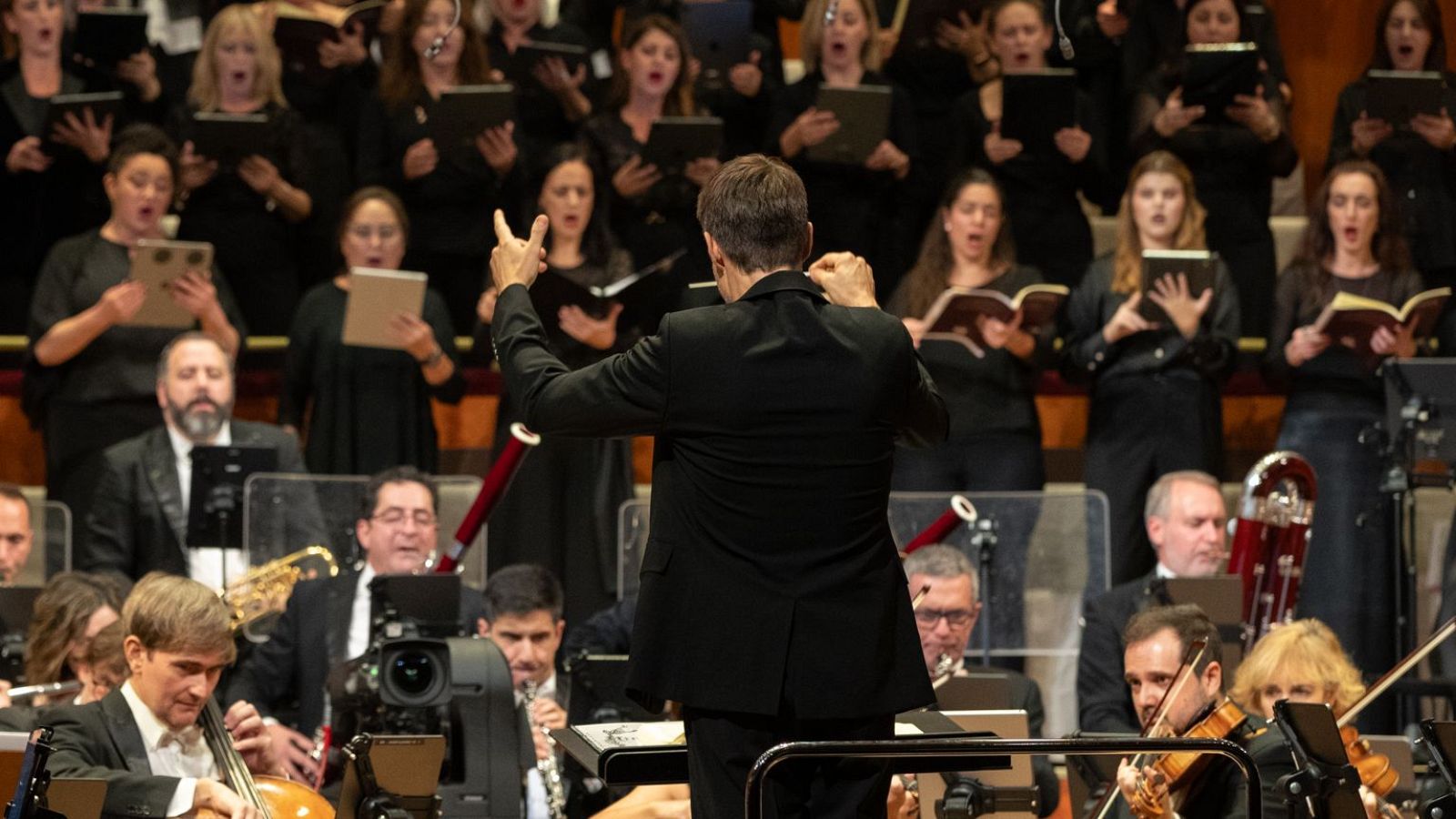 Una orquesta y un coro, ambos vestidos de negro, actúan juntos. El director, de espaldas a la cámara, guía a los músicos y cantantes. En la parte superior se lee el nombre de la agrupación.