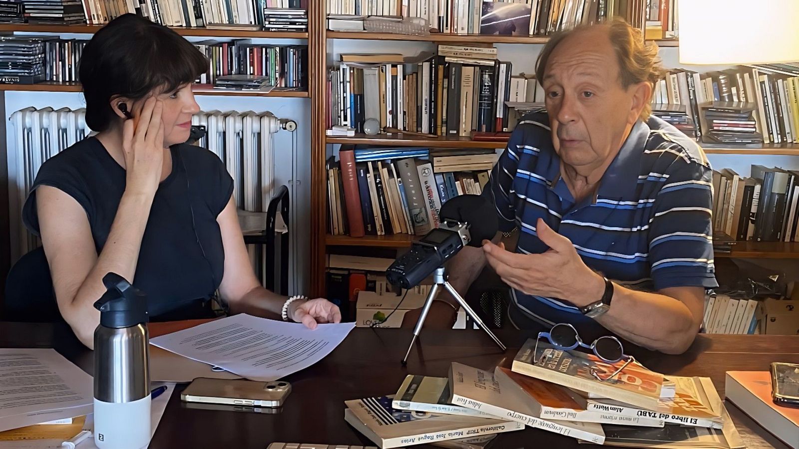 En un estudio, dos personas están sentadas frente a una mesa con un micrófono. Una de ellas lleva un auricular y una camiseta oscura, mientras que la otra gesticula con las manos. Hay libros y gafas sobre la mesa.