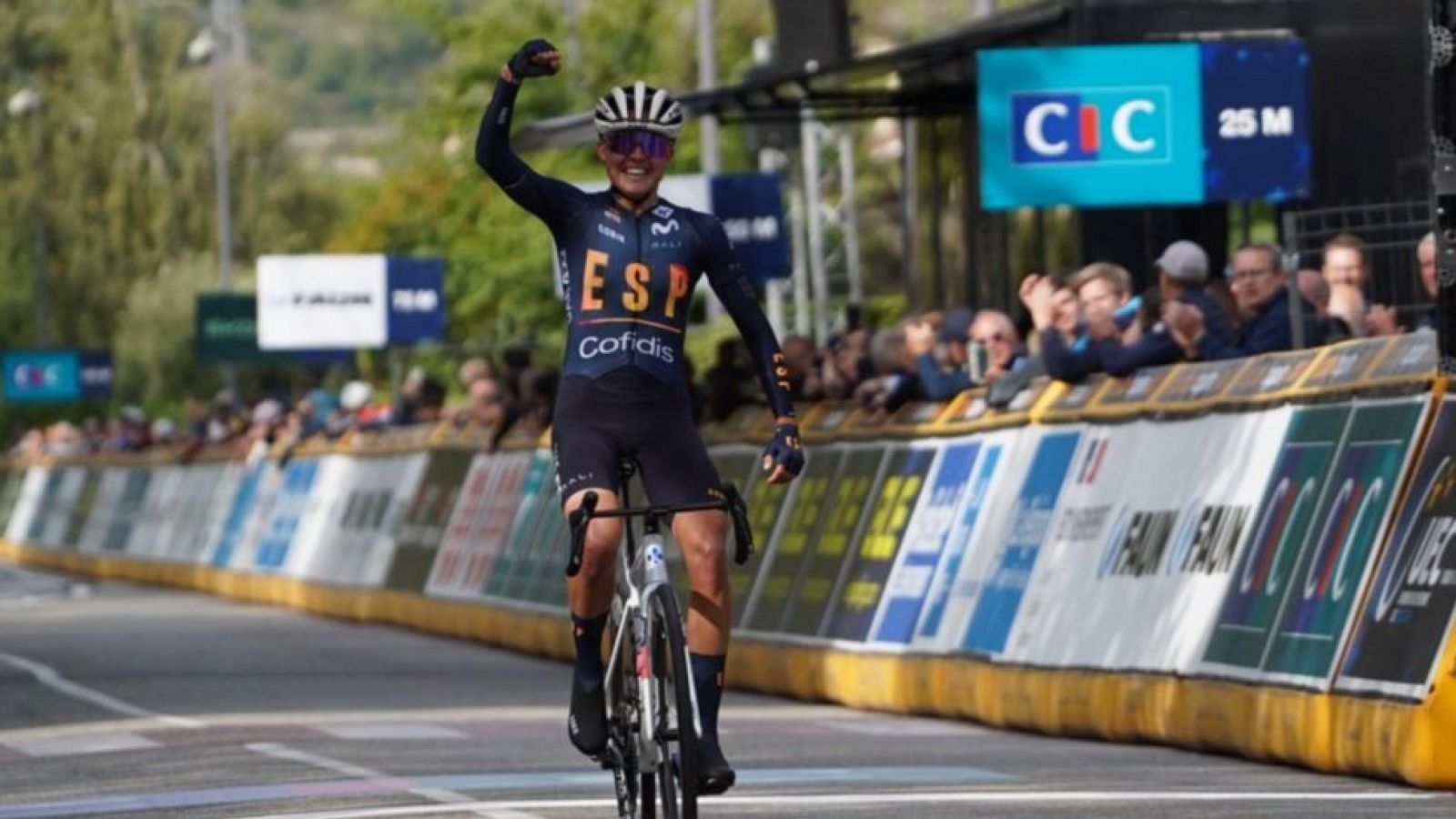 Una ciclista, vestida con un maillot azul oscuro y casco, celebra su victoria sobre la línea de meta, levantando el brazo derecho. Se encuentra en una carretera con espectadores a ambos lados.