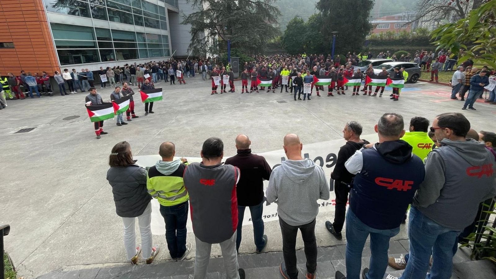 Una manifestación con banderas se desarrolla frente a un edificio moderno. Un grupo de personas con chalecos y banderas de Palestina lideran la protesta, seguidos por una multitud mayor.