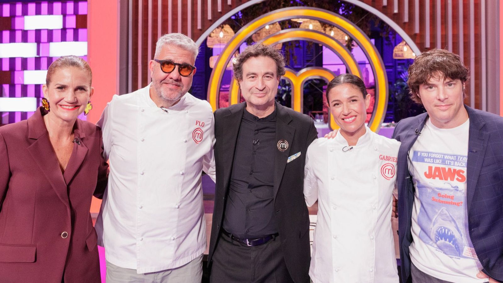 Un grupo de cinco personas posa frente al logo de un programa de cocina. Entre ellos, se observan individuos con chaquetas de chef y otros con vestimenta casual, como una camiseta con un logo.