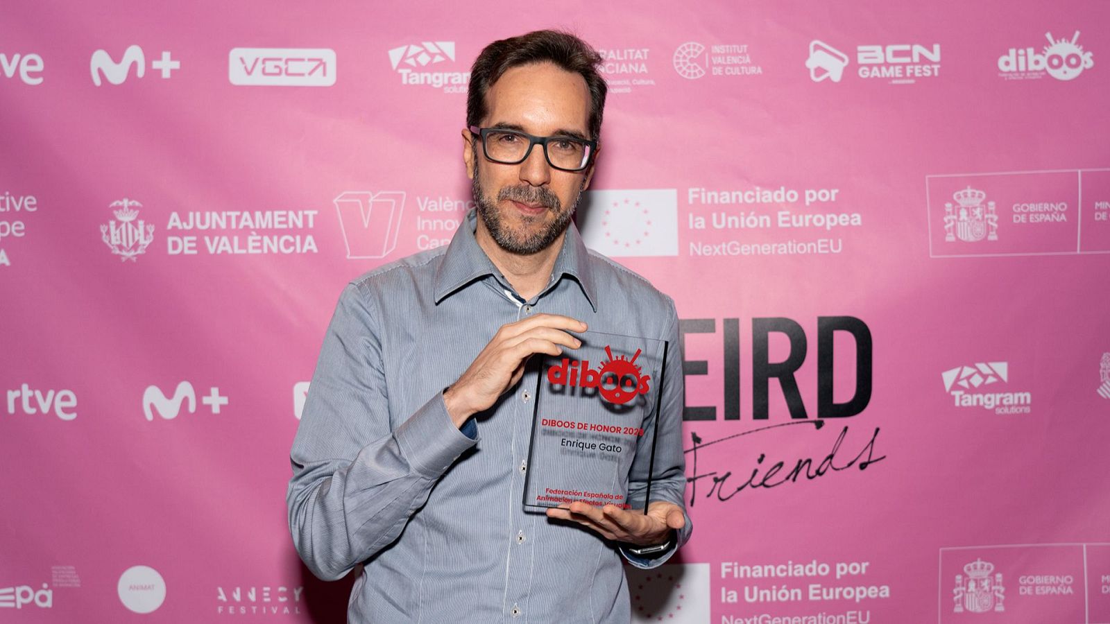 Enrique Gato, con gafas y camisa clara, recibe un premio transparente con un logotipo y una inscripción, posiblemente en un evento. El fondo es de color rosa y presenta varios logotipos.