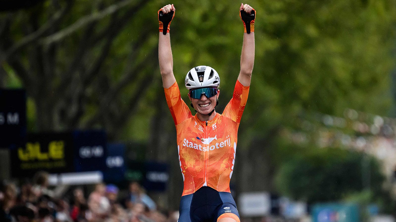 Demi Vollering, con un maillot naranja y gafas de sol, celebra su victoria con los brazos en alto y una sonrisa. El fondo muestra a la multitud y vegetación, en un evento deportivo de ciclismo.