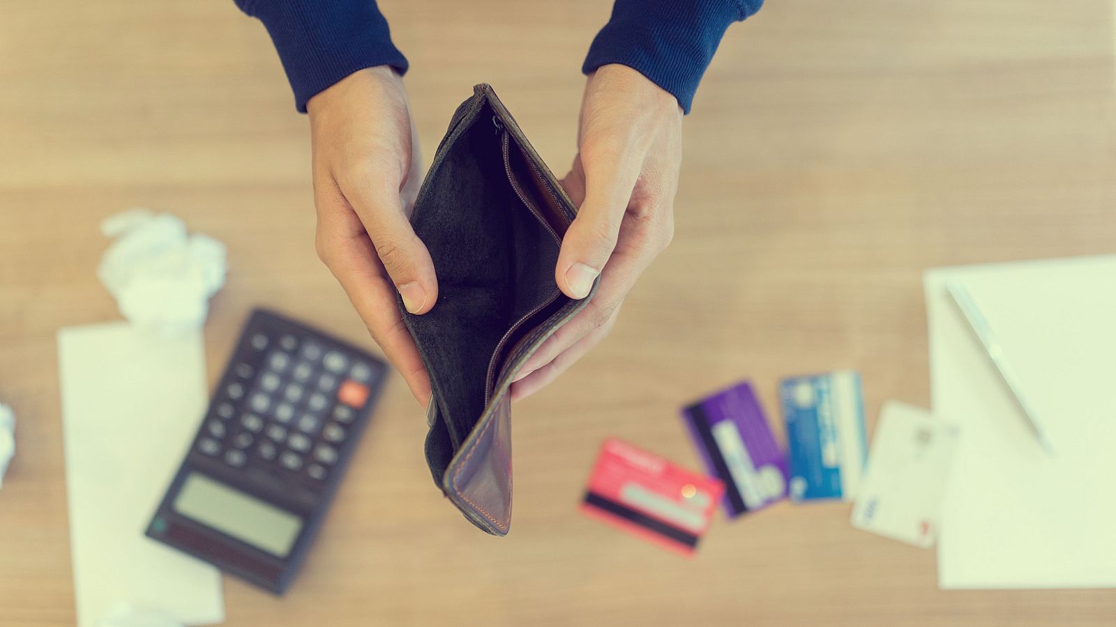 Una cartera de cuero marrón vacía es sostenida por manos, rodeada de objetos como una calculadora, papel arrugado, tarjetas de crédito y un bloc de notas, sugiriendo dificultades económicas.