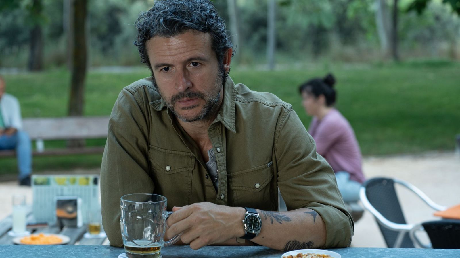 Diego Martín, con barba y pelo rizado, disfruta de una bebida y comida en una mesa al aire libre. Lleva una camisa verde oliva y un reloj de pulsera con correa negra, con un tatuaje en el brazo.