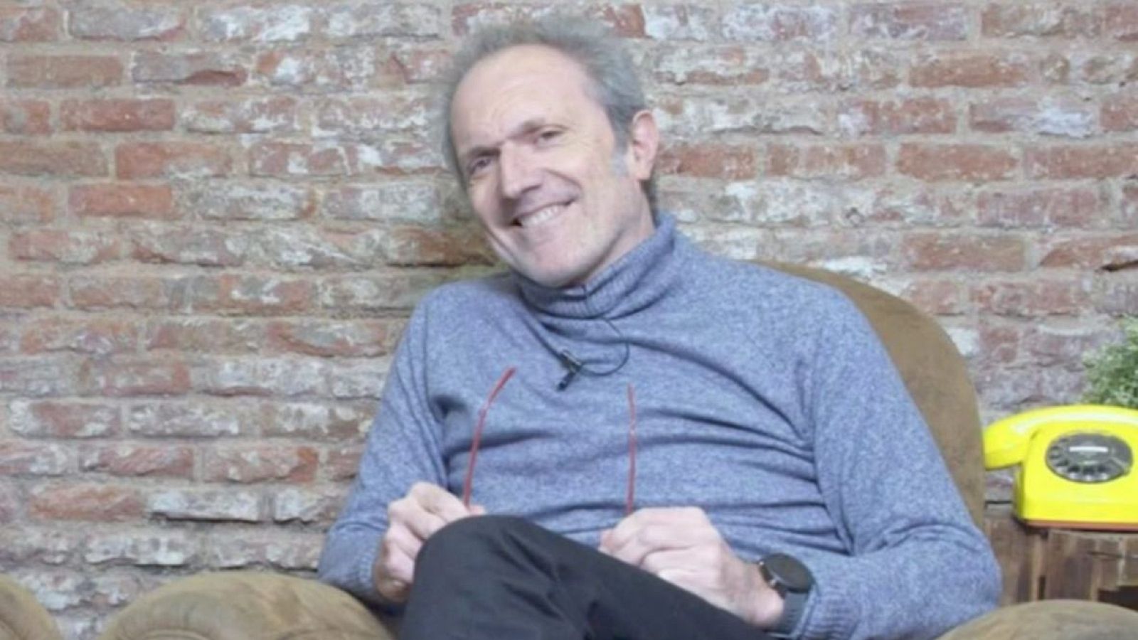 Carlos de la Cruz, con cabello canoso y una sonrisa, lleva un jersey azul grisáceo y pantalones oscuros. Sostiene unas gafas rojas y está sentado en un sillón. Detrás, una pared de ladrillo y un teléfono antiguo.