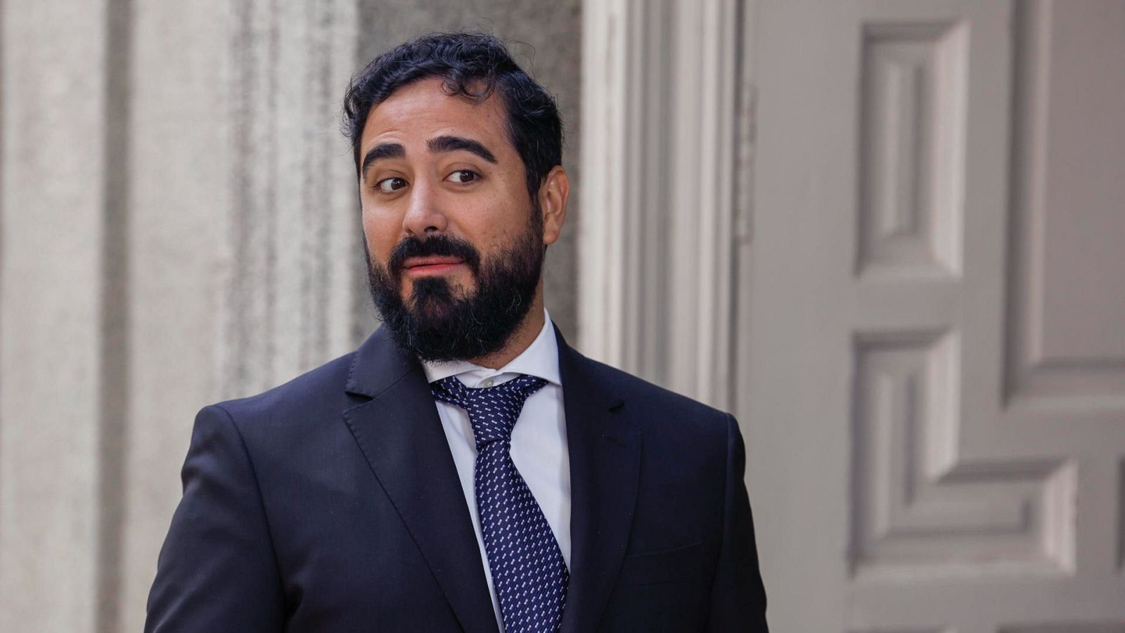 Alvise, en traje oscuro y corbata azul, llega al Tribunal Supremo.  Su expresión es seria; se encuentra frente a la entrada del edificio.