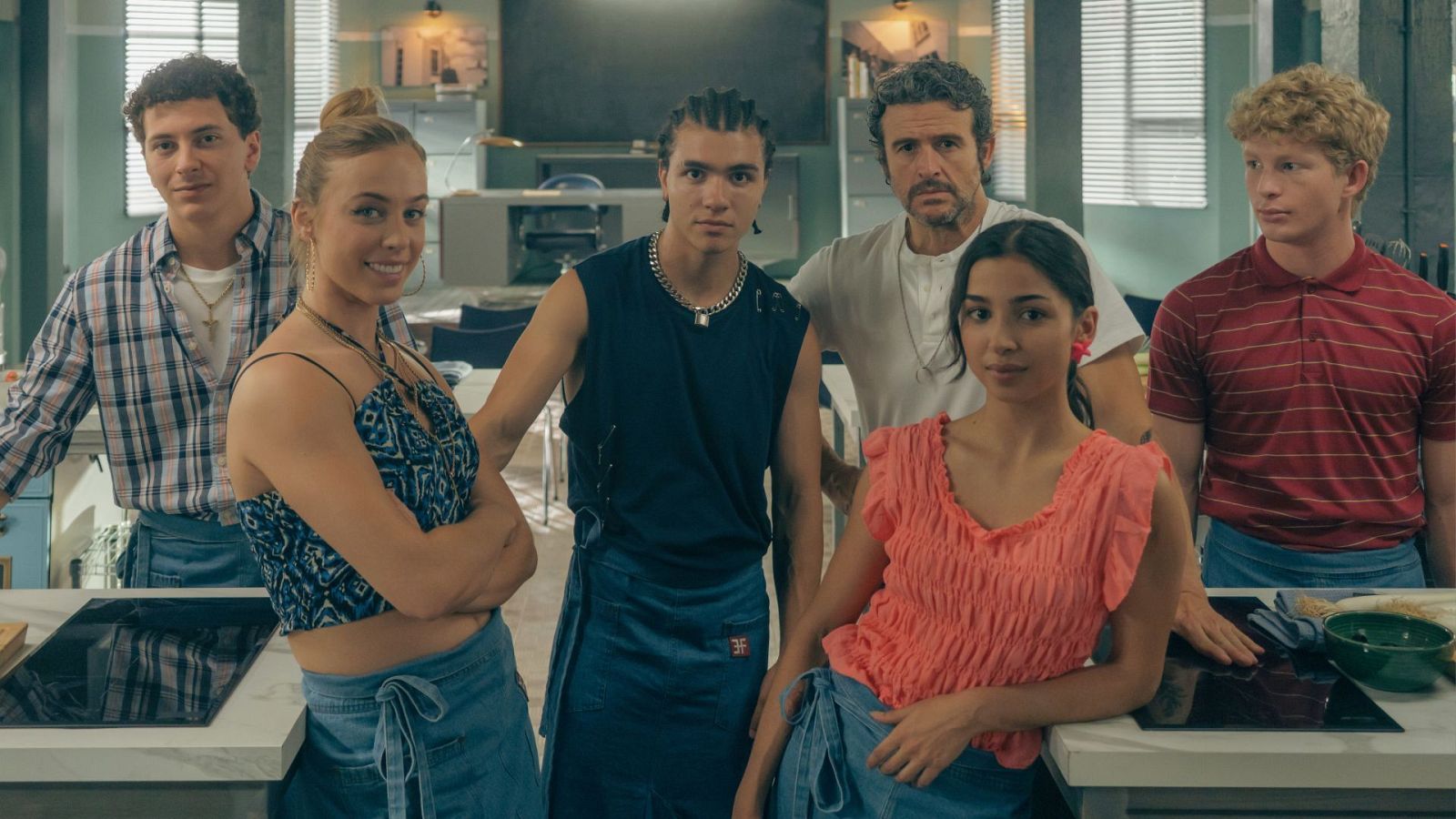 En una cocina, seis personas posan juntas. Se distinguen tres mujeres y tres hombres, con diferentes estilos de vestimenta, frente a una encimera con utensilios y una placa de cocina.