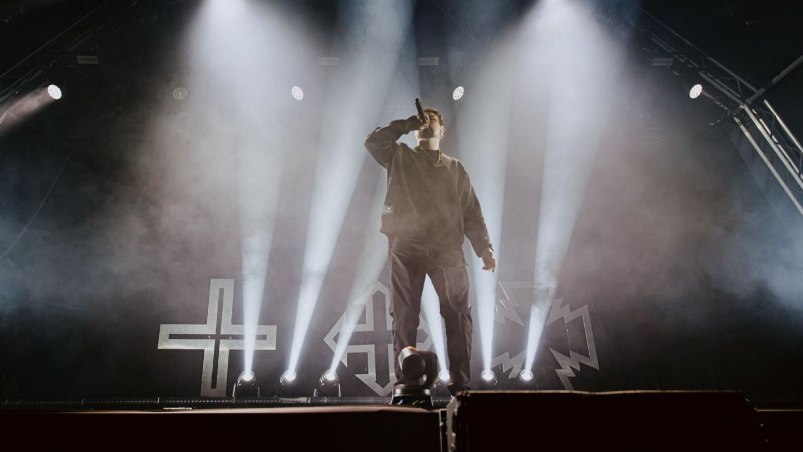 En un escenario iluminado, un hombre con vestimenta oscura canta con un micrófono en la mano, mientras levanta la otra. Detrás de él, se aprecia un logotipo con forma de cruz.