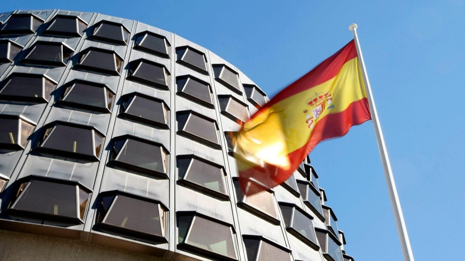 Edificio con fachada curva y metálica, ventanas trapezoidales y bandera española ondeando.
