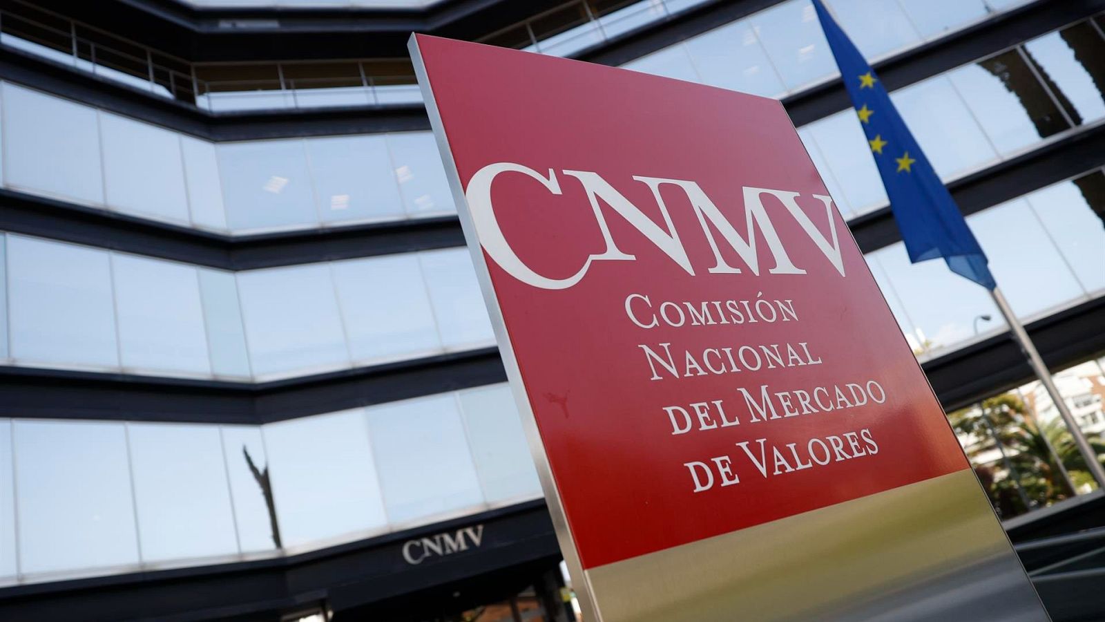 Cartel rojo 'CNMV' en edificio acristalado, con bandera europea cercana.  Probable sede de la Comisión Nacional del Mercado de Valores.
