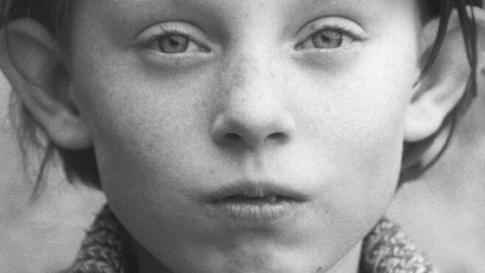 Primer plano en blanco y negro del rostro de un niño o niña, con ojos claros y labios finos, mirando directamente a la cámara. La imagen tiene una textura granulada.