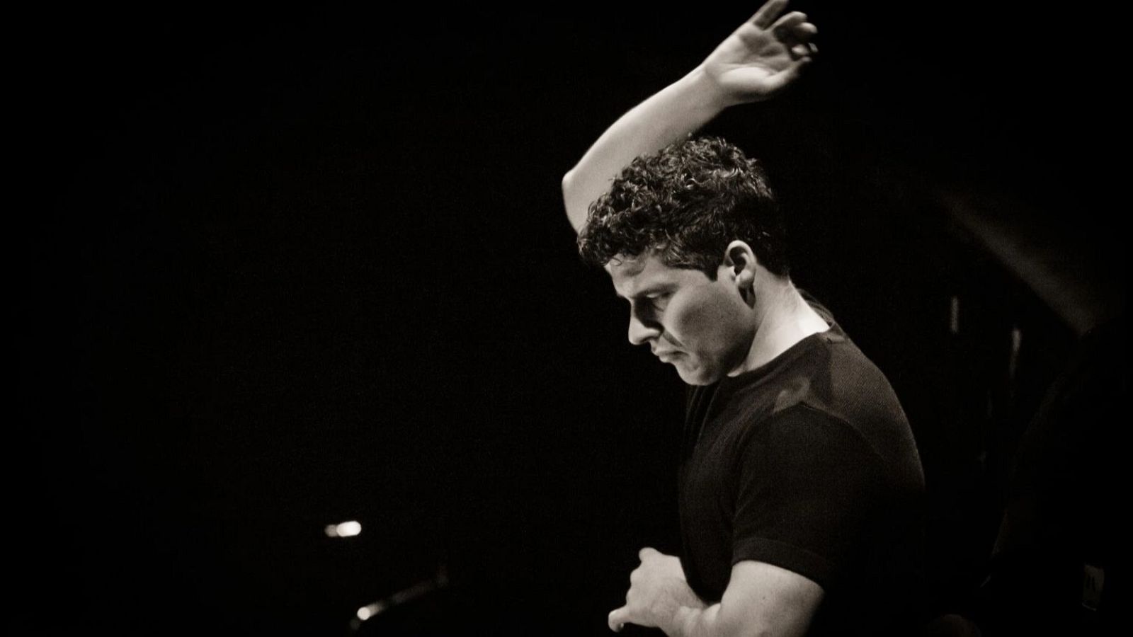 Retrato en blanco y negro de un hombre con cabello rizado y oscuro, de perfil, con un brazo levantado, posiblemente dirigiendo. Viste una camiseta negra y se encuentra en un entorno oscuro.