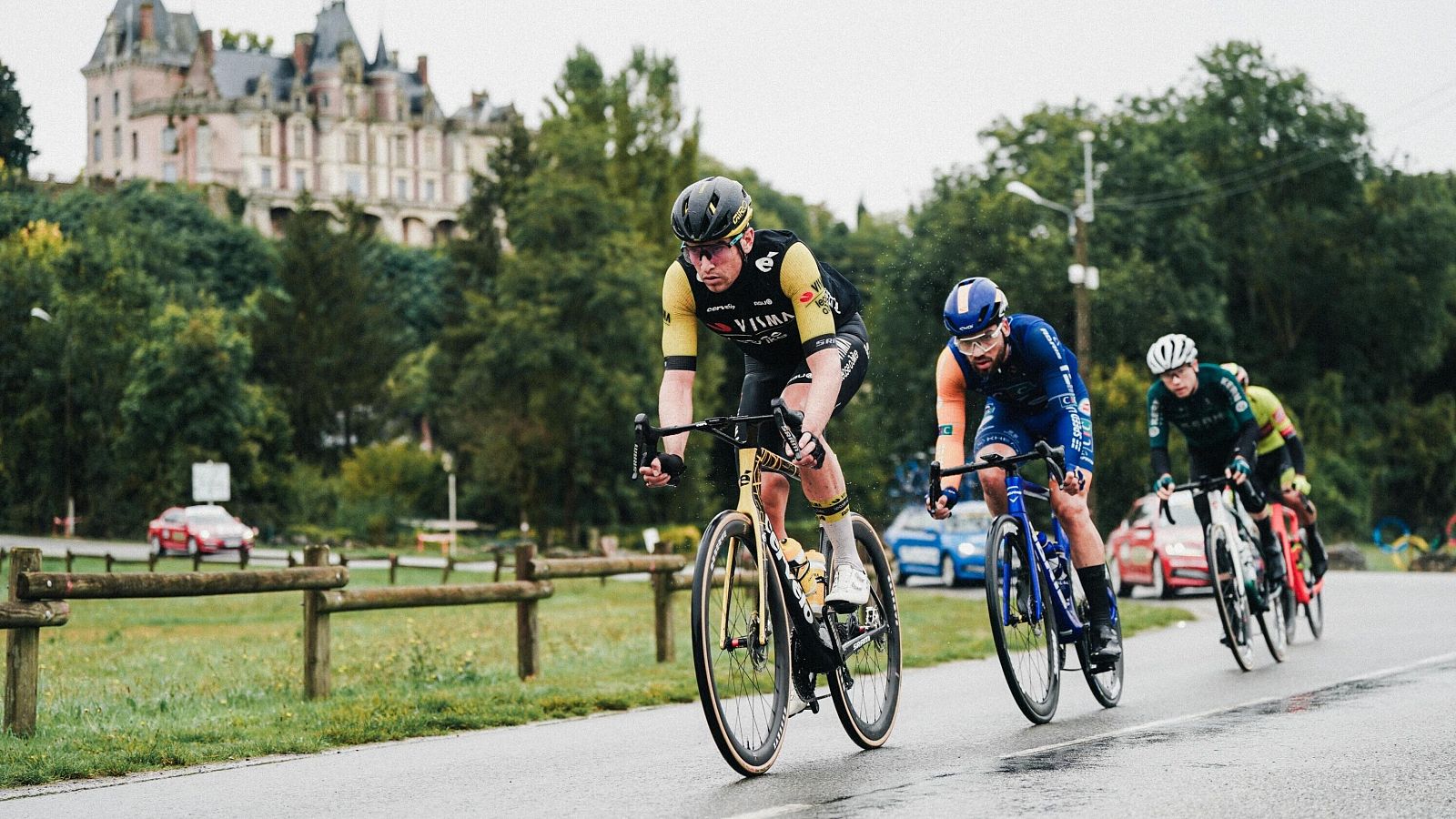 Tres ciclistas pedalean en una carretera asfaltada y mojada, durante una competición. Uno lleva un maillot negro con detalles amarillos, otro azul con detalles naranjas y el tercero verde oscuro. Un castillo se alza en el horizonte.