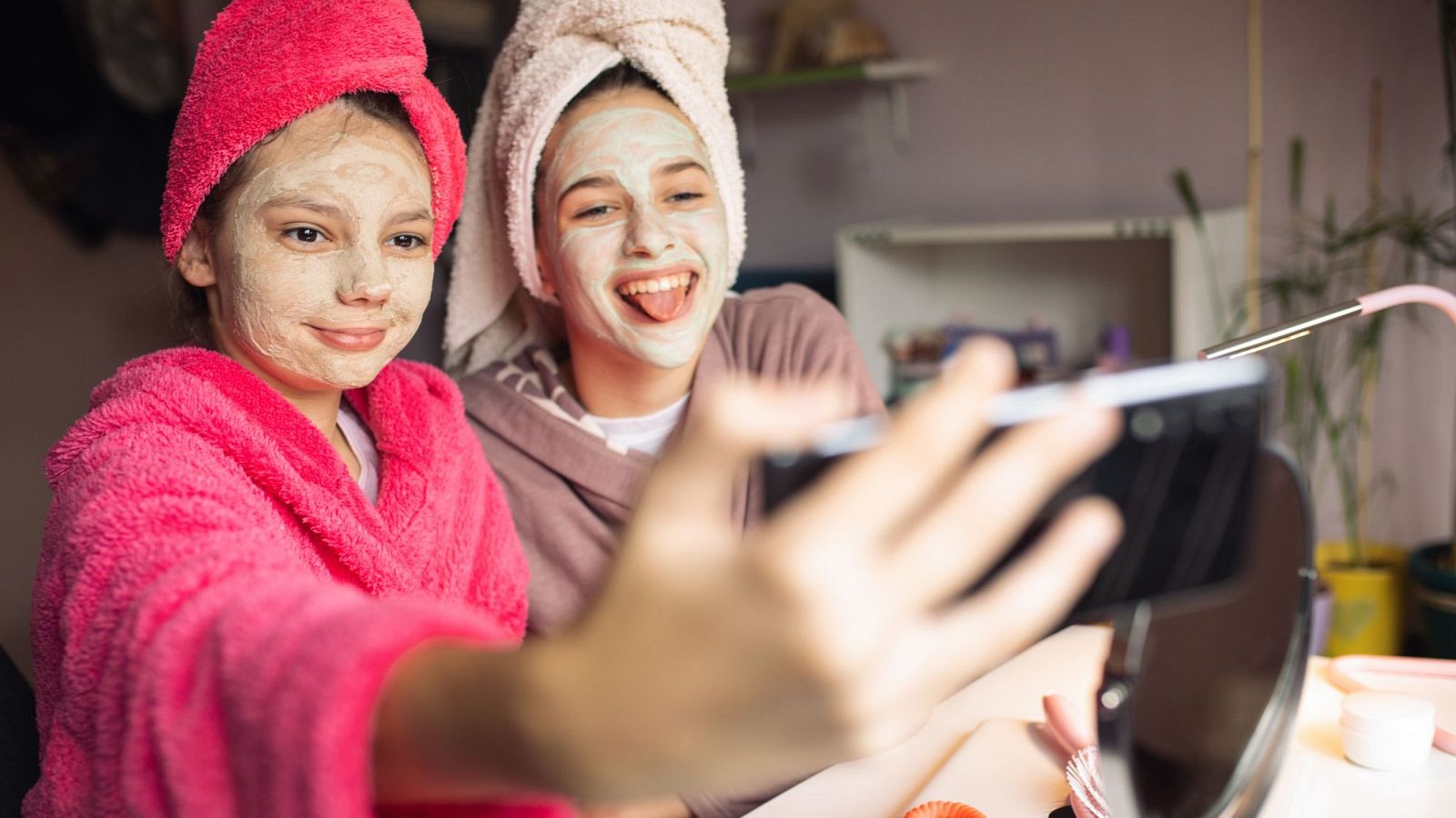 Dos chicas jóvenes, con mascarillas y toallas, posan con un teléfono móvil. Una lleva una bata rosa y sonríe, la otra una sudadera marrón y hace una mueca. En la mesa hay un espejo con luz y productos de belleza.