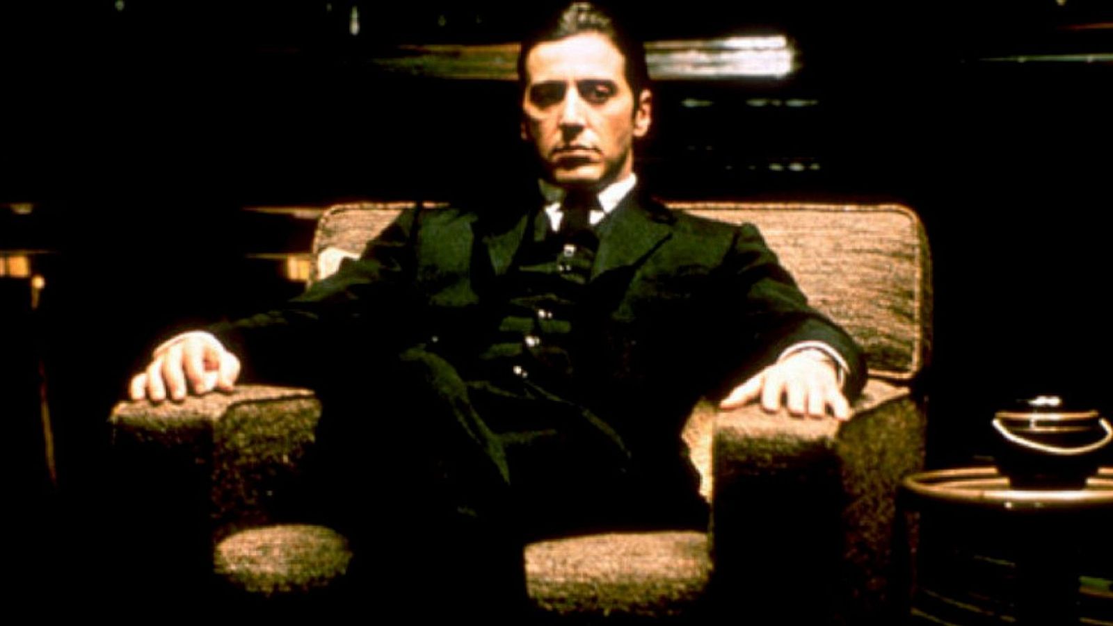 Al Pacino, como Michael Corleone, aparece sentado en un sillón, vestido con un traje oscuro y con una expresión intensa. La iluminación sombría crea un ambiente de poder y misterio.