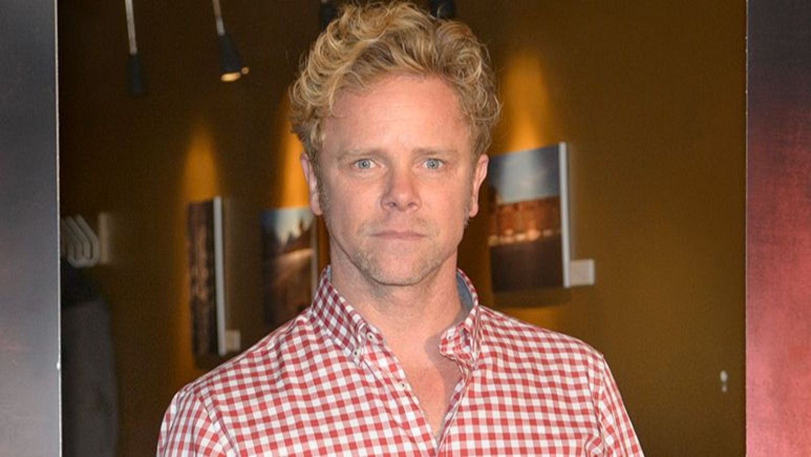 Un hombre de piel clara, cabello rubio rizado y ojos azules, identificado como Jimmy Shaw, posa con una camisa de cuadros rojos y blancos frente a una pared marrón con fotos y luces.