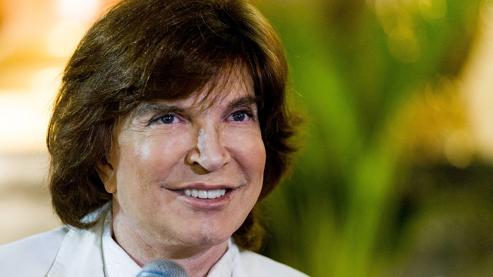 Primer plano de Camilo Sesto, con cabello castaño oscuro y flequillo, sonriendo levemente. Viste camisa blanca y un micrófono se encuentra en la imagen, con un fondo difuminado en tonos verdes y amarillos.