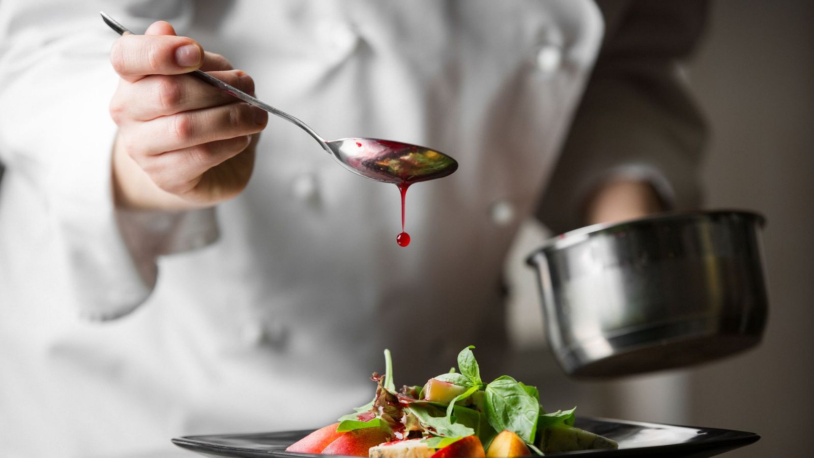 Un chef, con chaqueta blanca, adereza un plato con una cuchara, dejando caer salsa roja sobre una ensalada con hojas verdes y fruta. A la derecha, un cazo de metal.