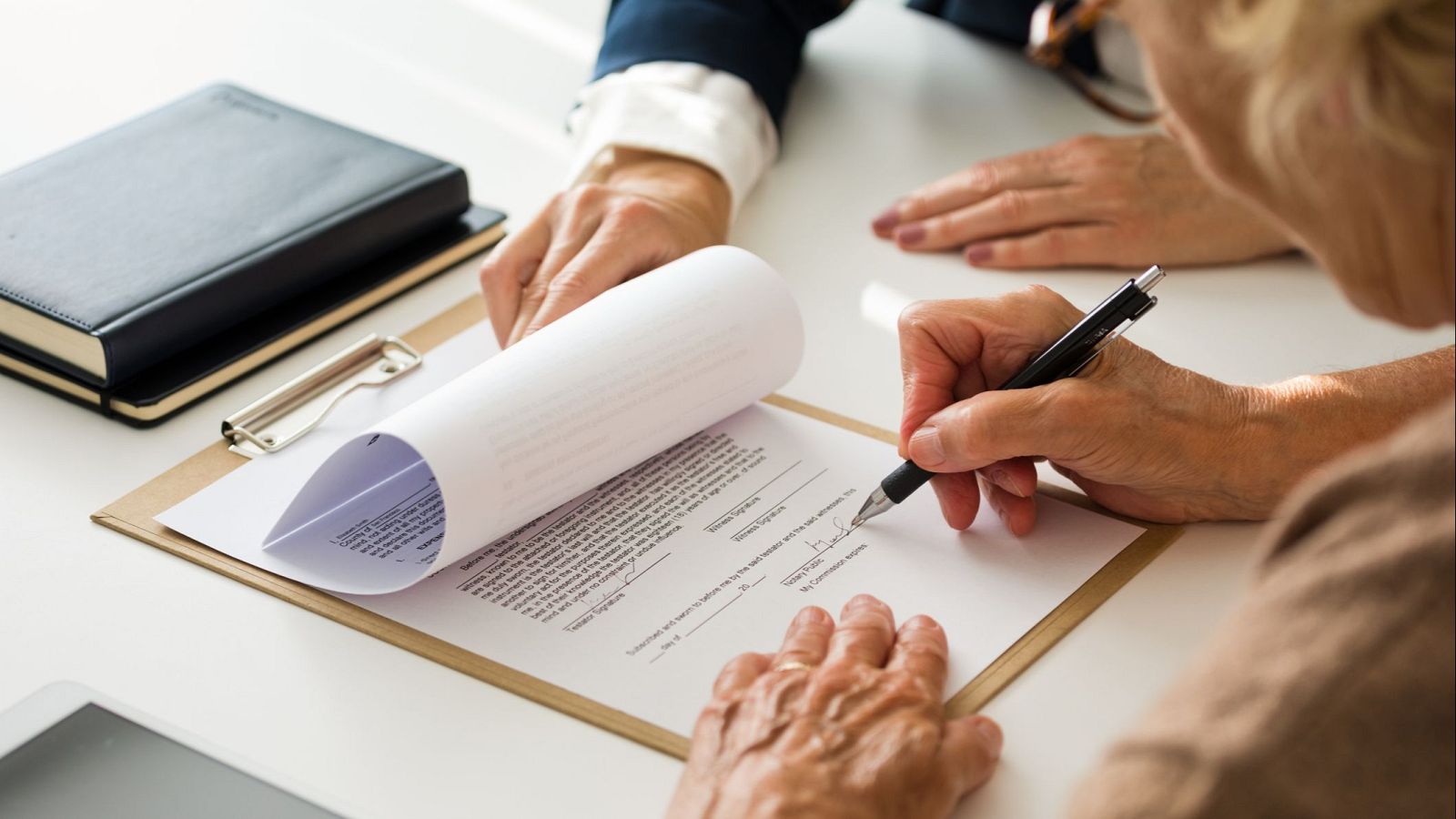 Una persona mayor, vestida con una camisa clara, firma un documento con un bolígrafo negro, mientras otra persona, con una chaqueta azul, observa. En la mesa hay una tablet y cuadernos.