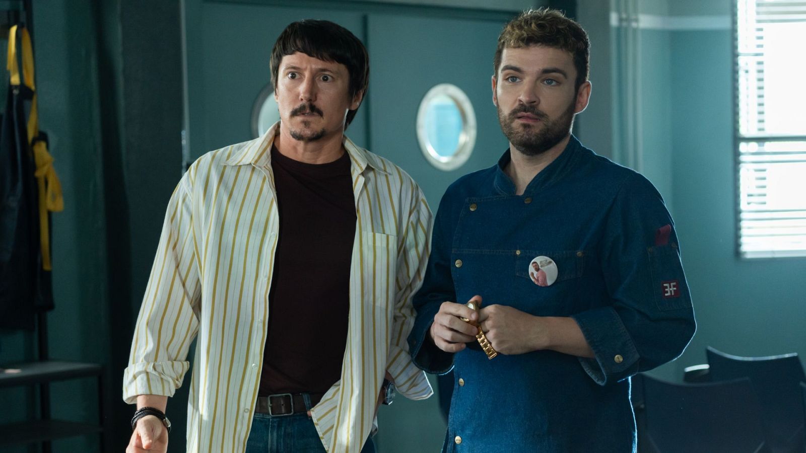 Dos hombres en una cocina: uno con camisa a rayas y vaqueros, el otro con chaqueta de chef azul, pin, pelo rizado y barba. Se observa una puerta con ventanas circulares y una ventana con persianas.