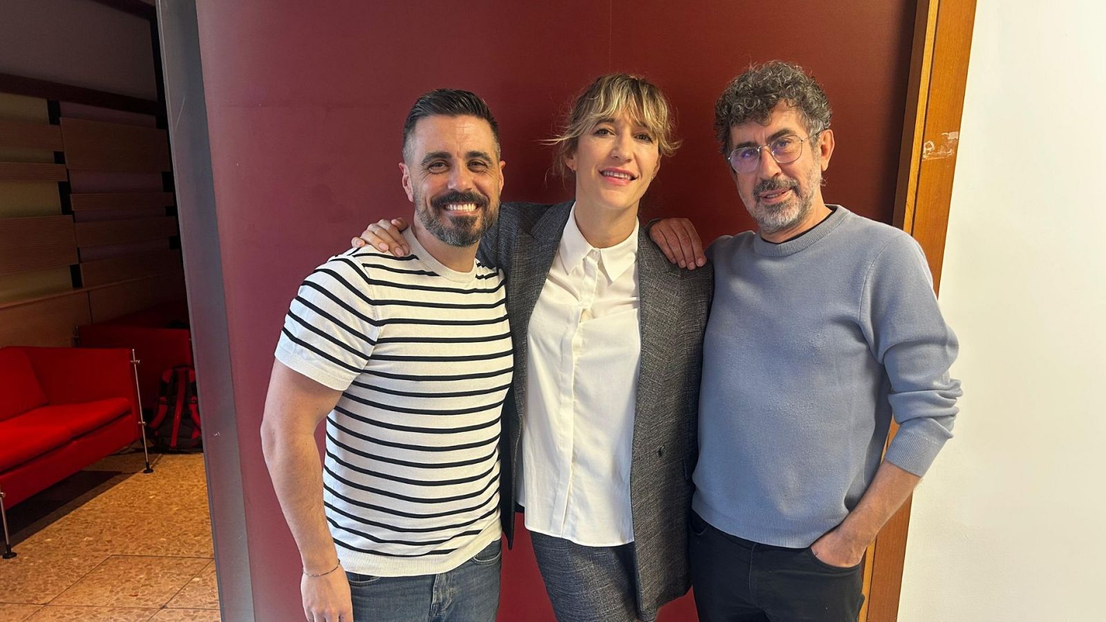 Tres personas sonríen a la cámara. Un hombre con barba y camiseta a rayas, una mujer con chaqueta gris y un hombre con gafas y jersey azul, posan frente a una pared roja con una sección blanca y un sofá rojo.