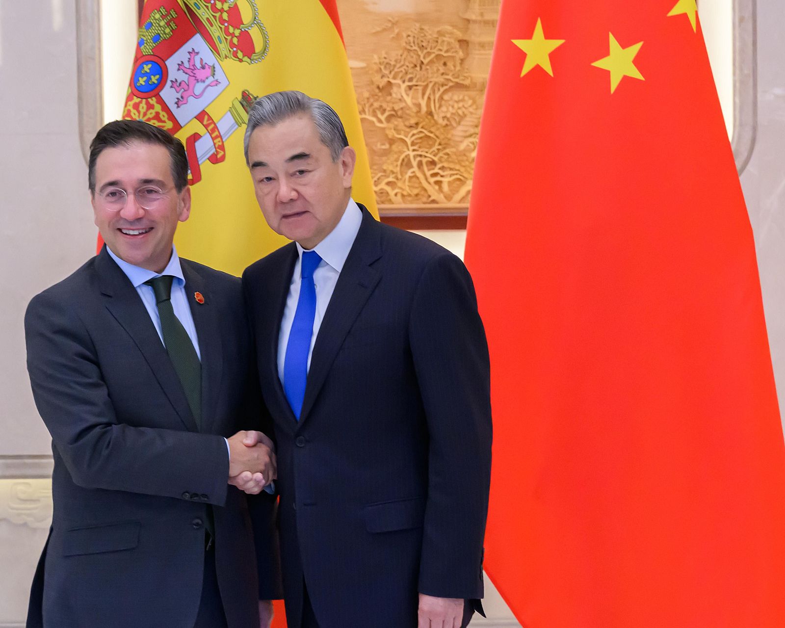 Dos ministros, uno español y otro chino, se dan la mano sonriendo. El ministro español viste un traje oscuro con corbata verde, mientras que el ministro chino lleva un traje azul oscuro con corbata azul, frente a las banderas de sus países.